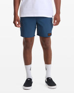 A person in a white T-shirt, Volcom Men's Truly Lib Work Trunk 17 blue shorts with a patch on the left leg, white logo socks, and black slip-on shoes stands against a plain white background.
