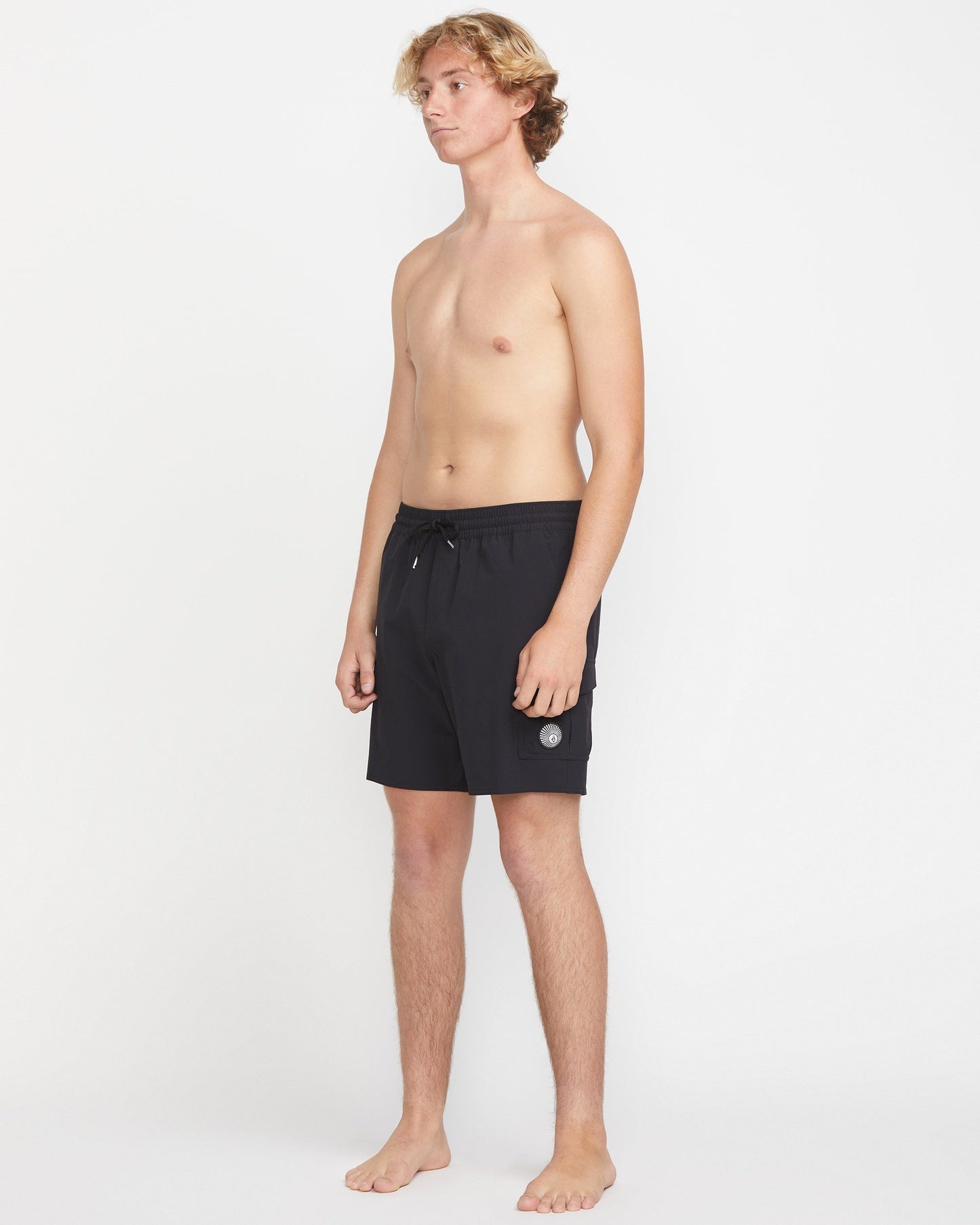 A young man with light skin and blonde hair stands barefoot against a white background, wearing Volcom Men's Truly Liberators 17" black trunk shorts with a circular logo and left-leg cargo pockets. He is shirtless and looking slightly to the side.