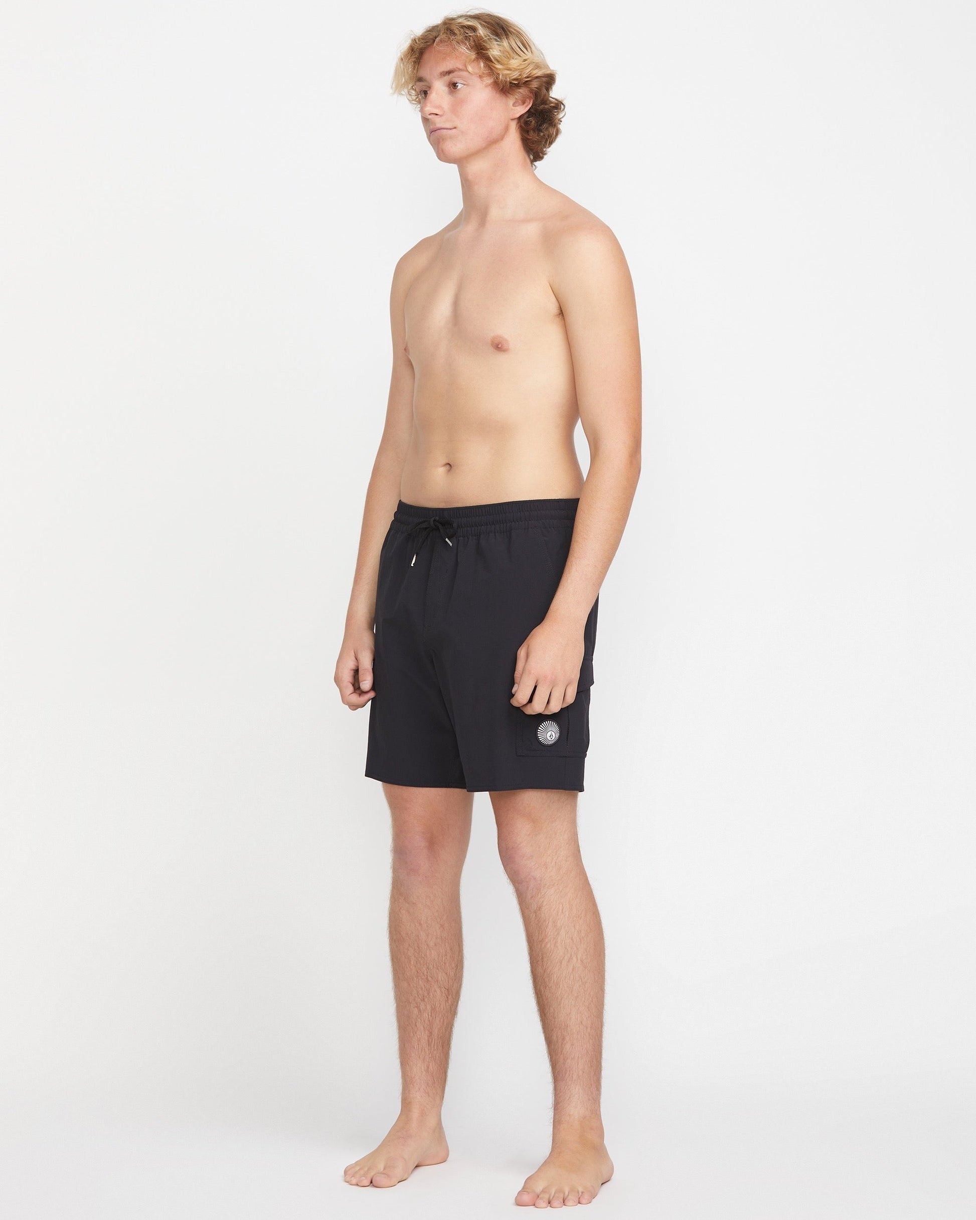 A young man with light skin and blonde hair stands barefoot against a white background, wearing Volcom Men's Truly Liberators 17" black trunk shorts with a circular logo and left-leg cargo pockets. He is shirtless and looking slightly to the side.