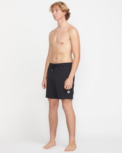 A young man with light skin and blonde hair stands barefoot against a white background, wearing Volcom Men's Truly Liberators 17" black trunk shorts with a circular logo and left-leg cargo pockets. He is shirtless and looking slightly to the side.