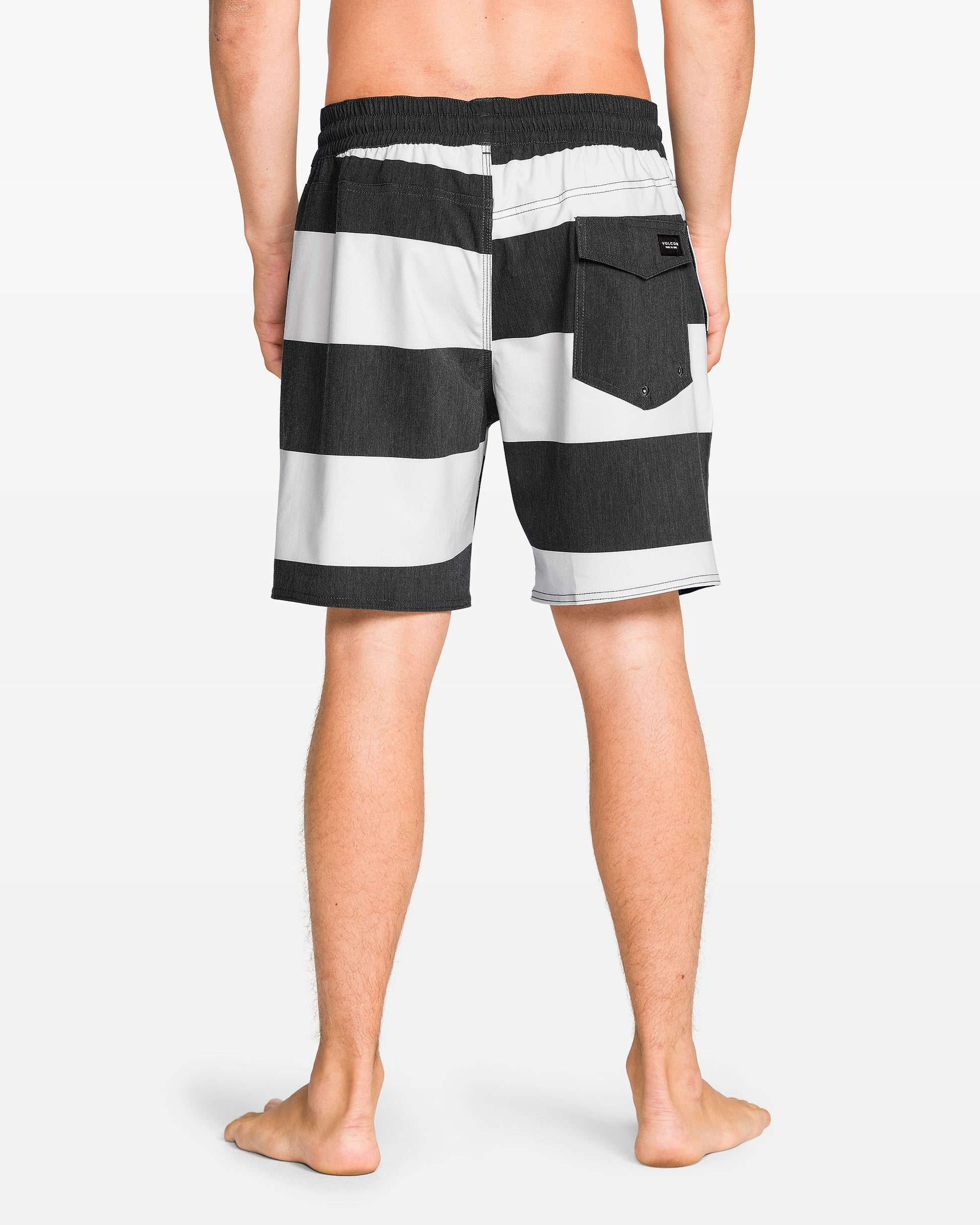 A man stands barefoot, facing away, wearing Volcom Men's Psyche Cotton 17" Trunk—black and white horizontal stripes, elastic waistband, back pocket.