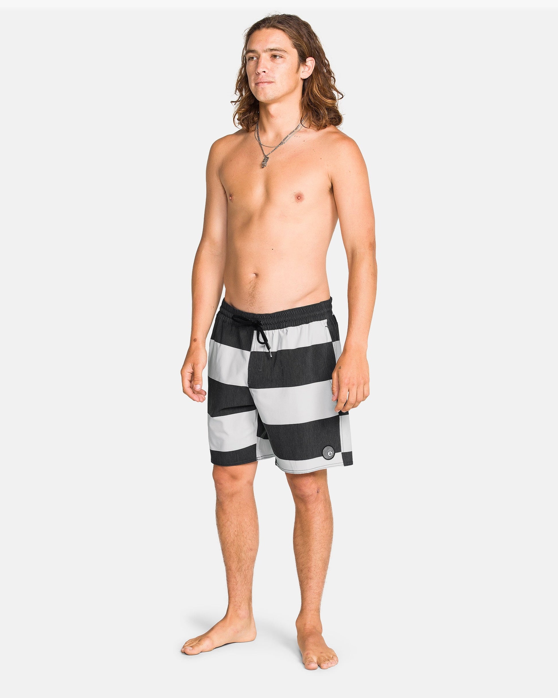 A young man with long brown hair stands barefoot on a white background, wearing Volcom Men's Psyche Cotton 17" Trunk swimwear and a necklace. He is shirtless and looking slightly to the side.
