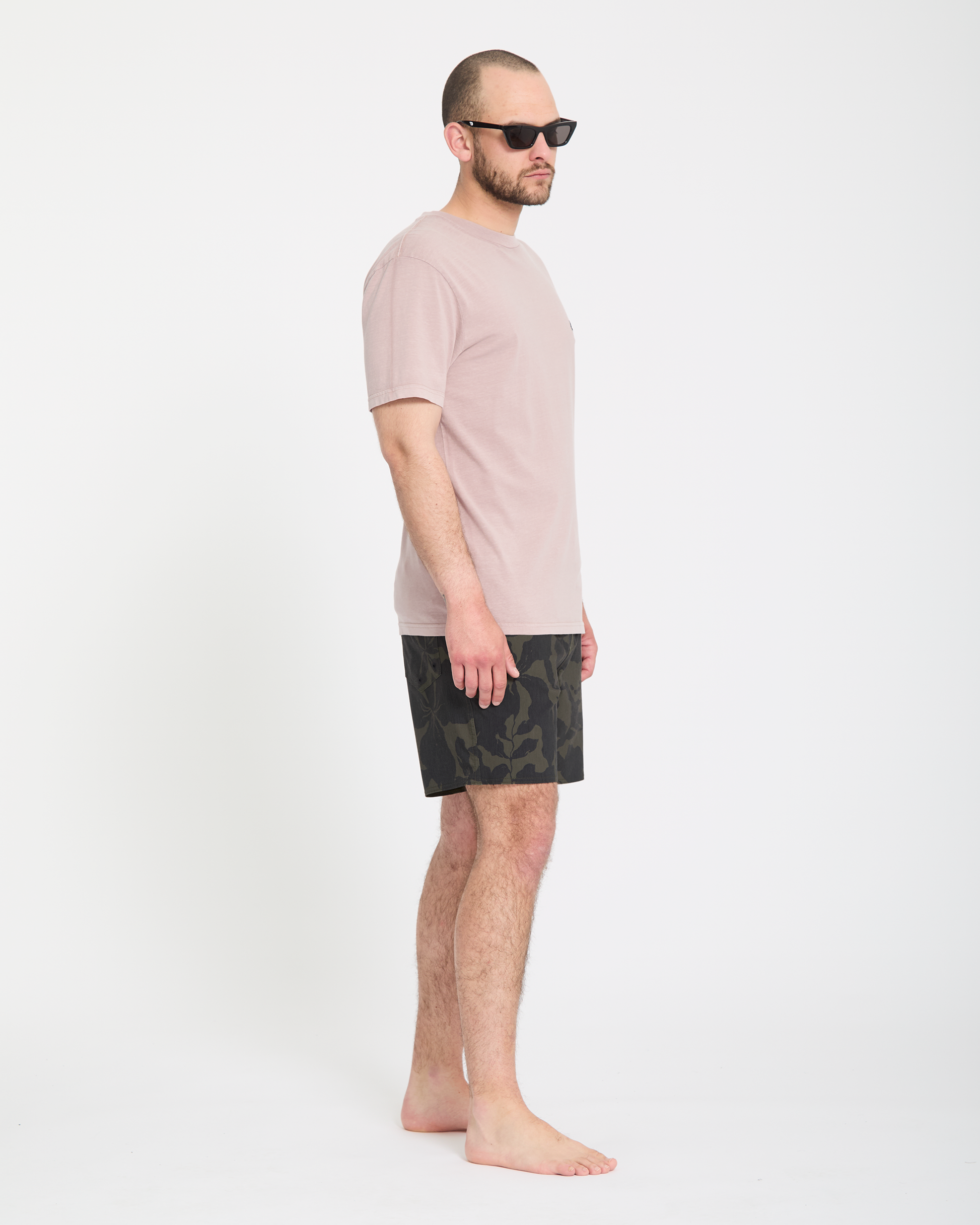 A man stands barefoot in profile against a white background, wearing black sunglasses, a pale pink t-shirt, and Volcom Men's Psyche Cotton Trunk 17 shorts.