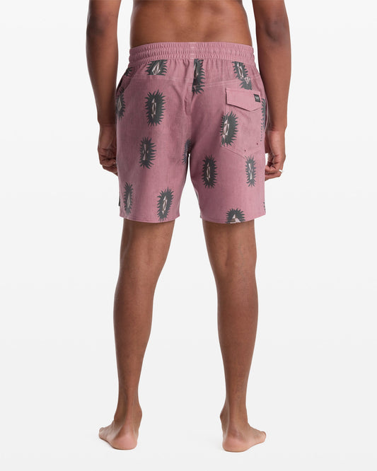 A person stands barefoot facing away, wearing Volcom Men's Psyche Cotton Trunk 17 shorts in pink with a black cactus pattern, featuring an elastic waistband and a back pocket.