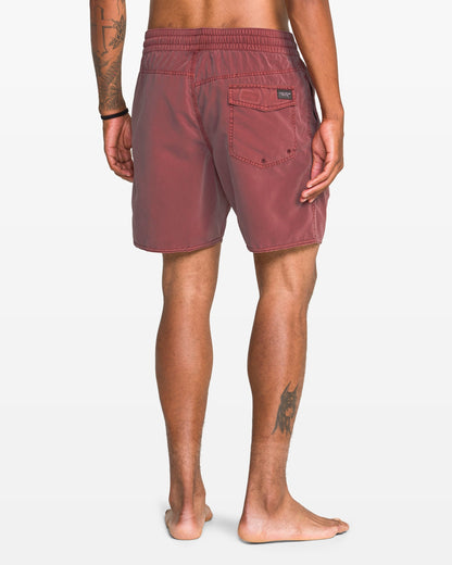 A barefoot person faces away in Volcom Men's Center Elastic Waist 17" Trunk—red, recycled polyester shorts with a back pocket. Tattoos on their left arm and right calf are visible against a plain white background.