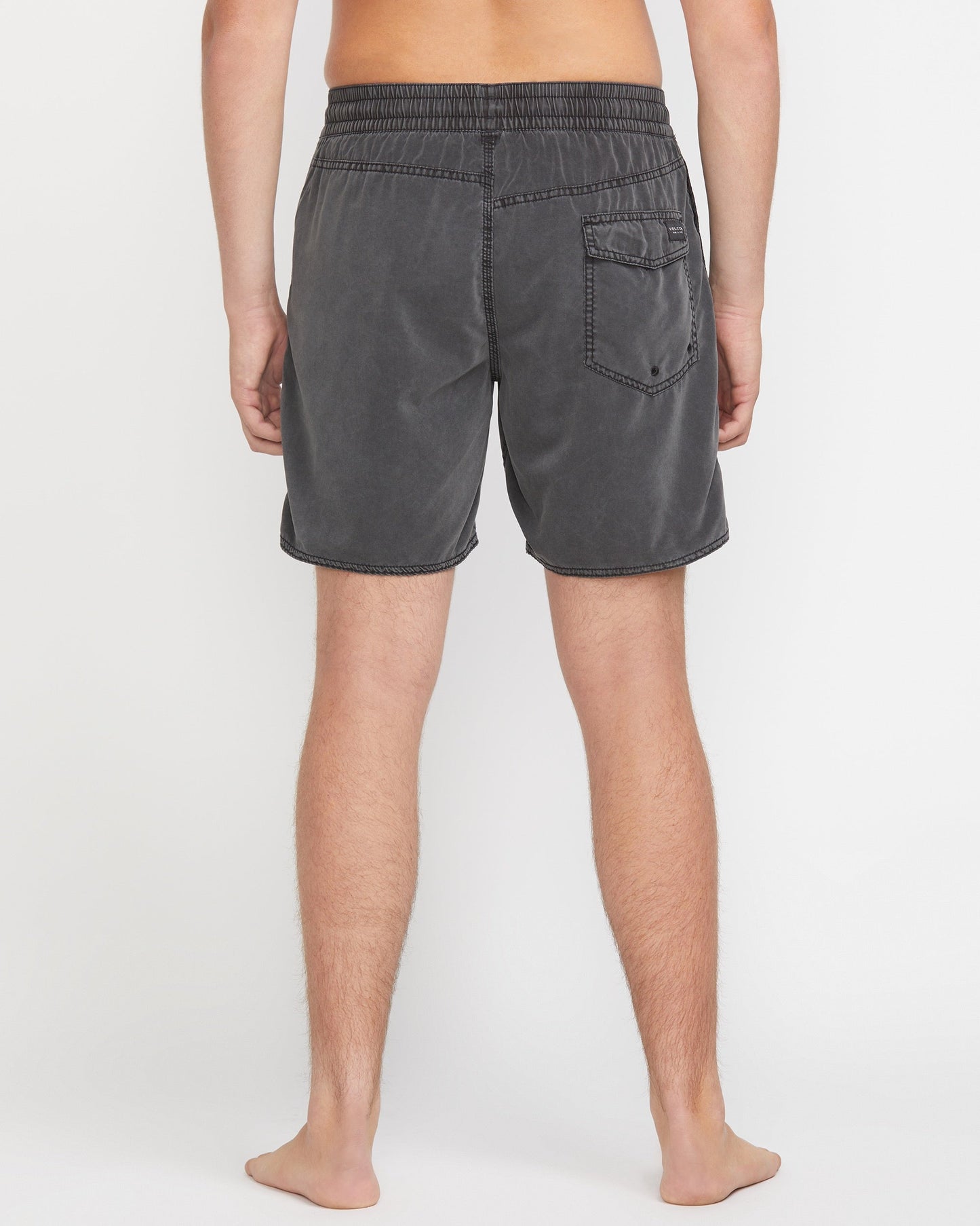 A person stands barefoot facing away, wearing Volcom Men's Center Elastic Waist 17" Trunk in charcoal gray with a single back pocket and mechanical stretch; the background is plain white.