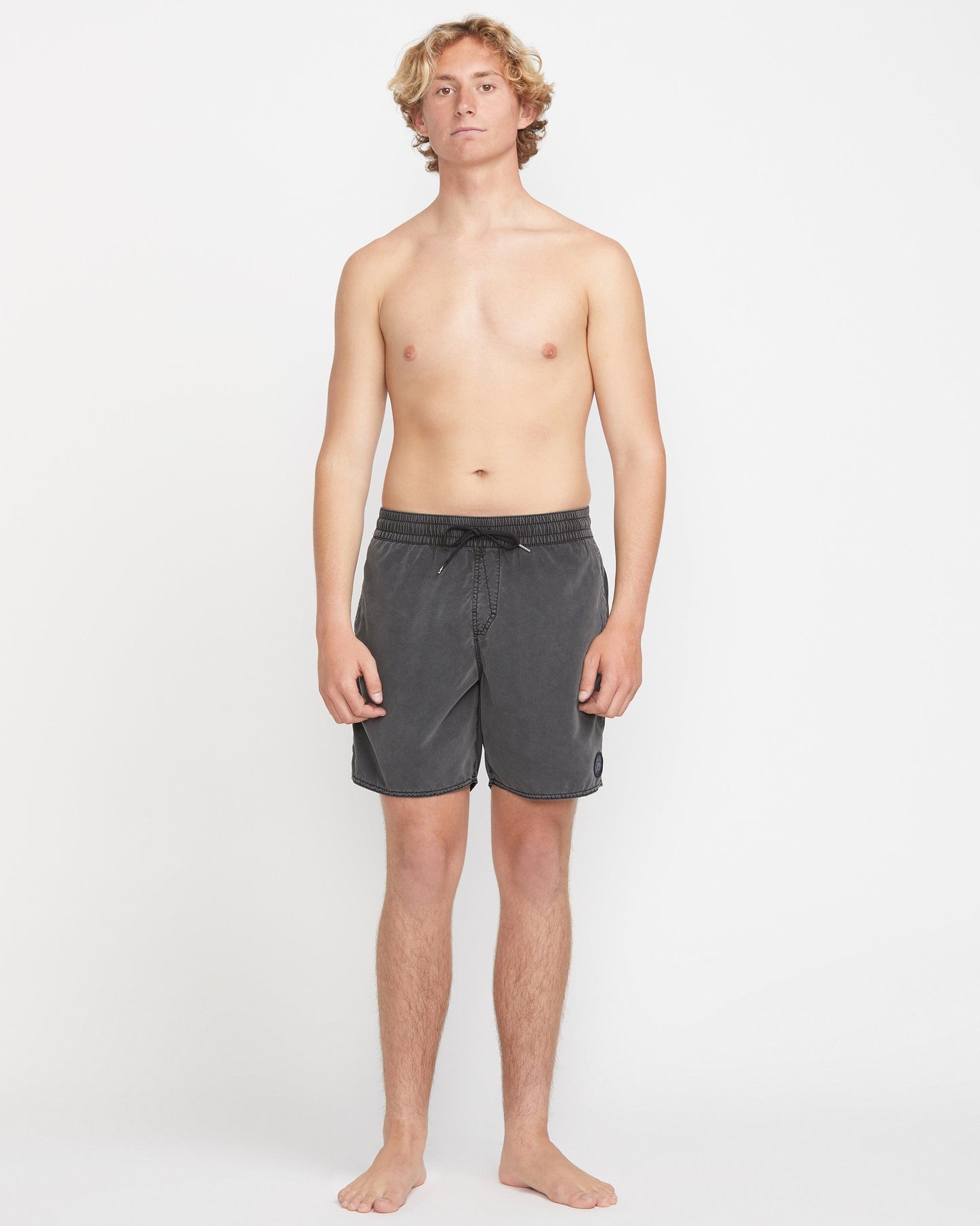 A young man with curly blond hair stands barefoot against a plain white background, wearing Volcom Men's Center Elastic Waist 17" Trunk in dark gray made from recycled polyester with mechanical stretch, facing forward neutrally with arms at his sides.