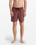 A person stands barefoot, facing forward against a white background, wearing Volcom Men's Center Elastic Waist Trunks in maroon with a black drawstring, elastic waist, and a small patch on the left leg.