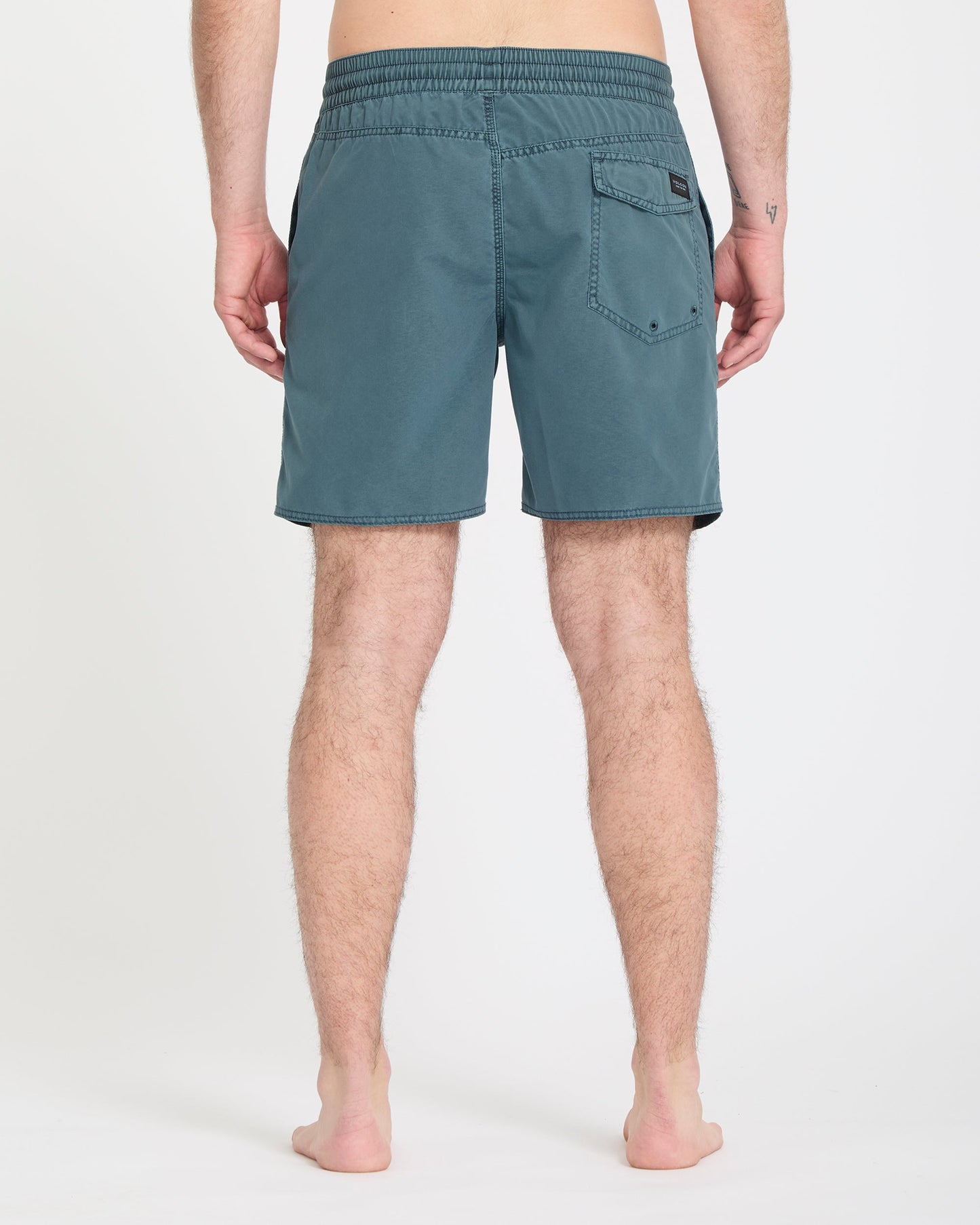 A person stands barefoot on a white background, shown from the back, wearing Volcom Men's Center Trunks 17 in teal with an elastic waistband and a single back pocket.