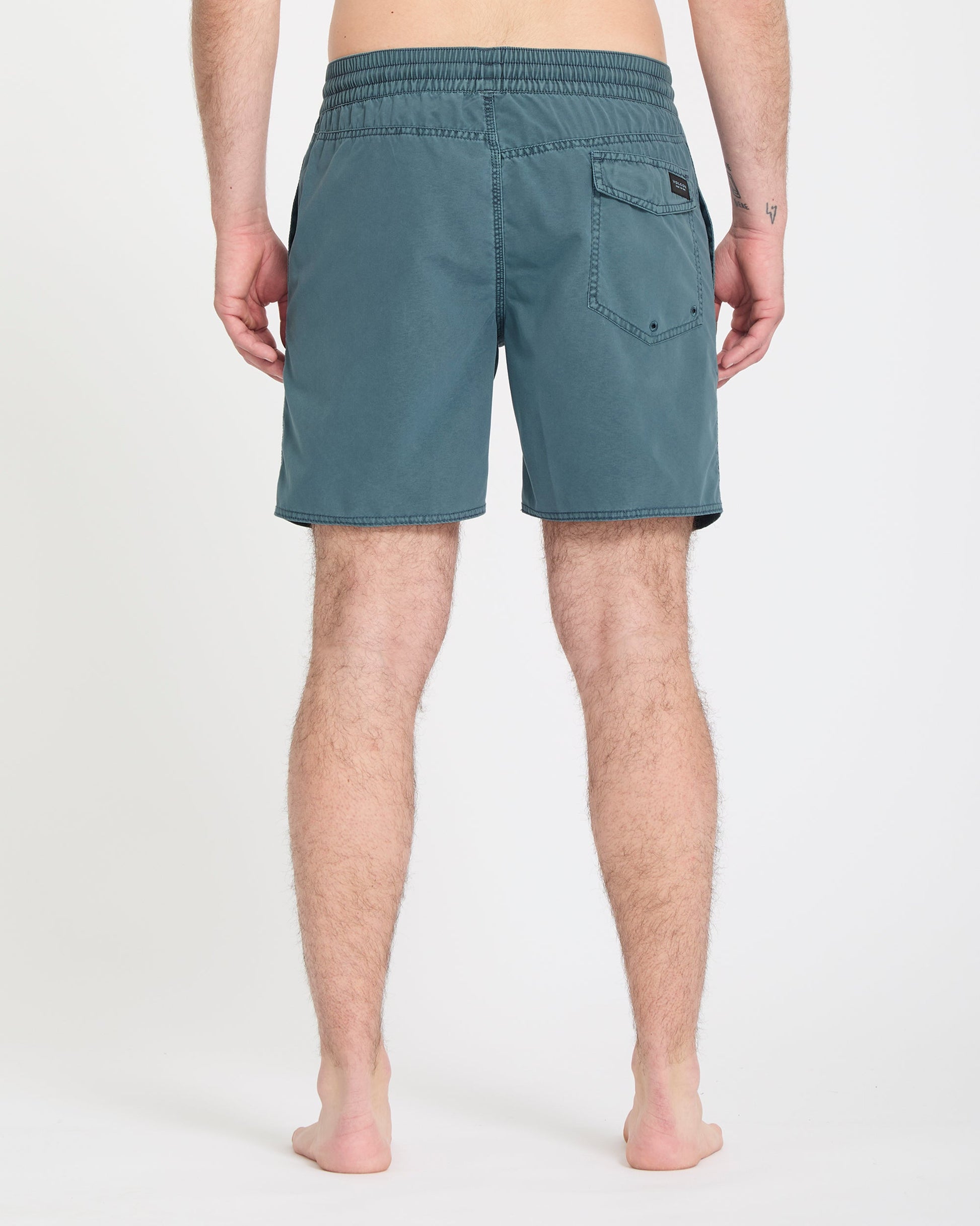 A person stands barefoot on a white background, shown from the back, wearing Volcom Men's Center Trunks 17 in teal with an elastic waistband and a single back pocket.