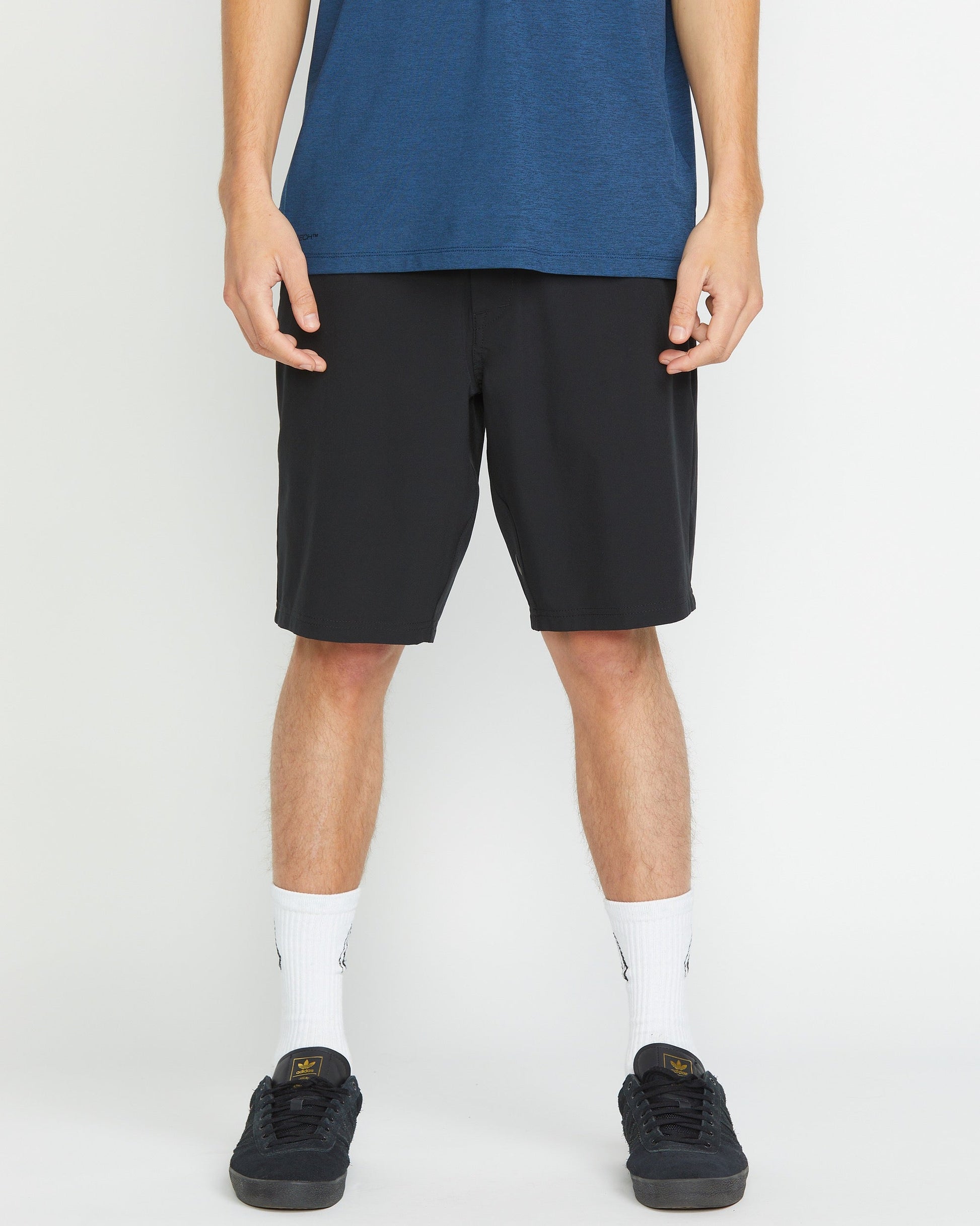 A person stands against a plain background, visible from shoulders down, wearing a blue T-shirt, Volcom Men's Frickin Cross Shred 20" Shorts in black, white crew socks, and black sneakers.