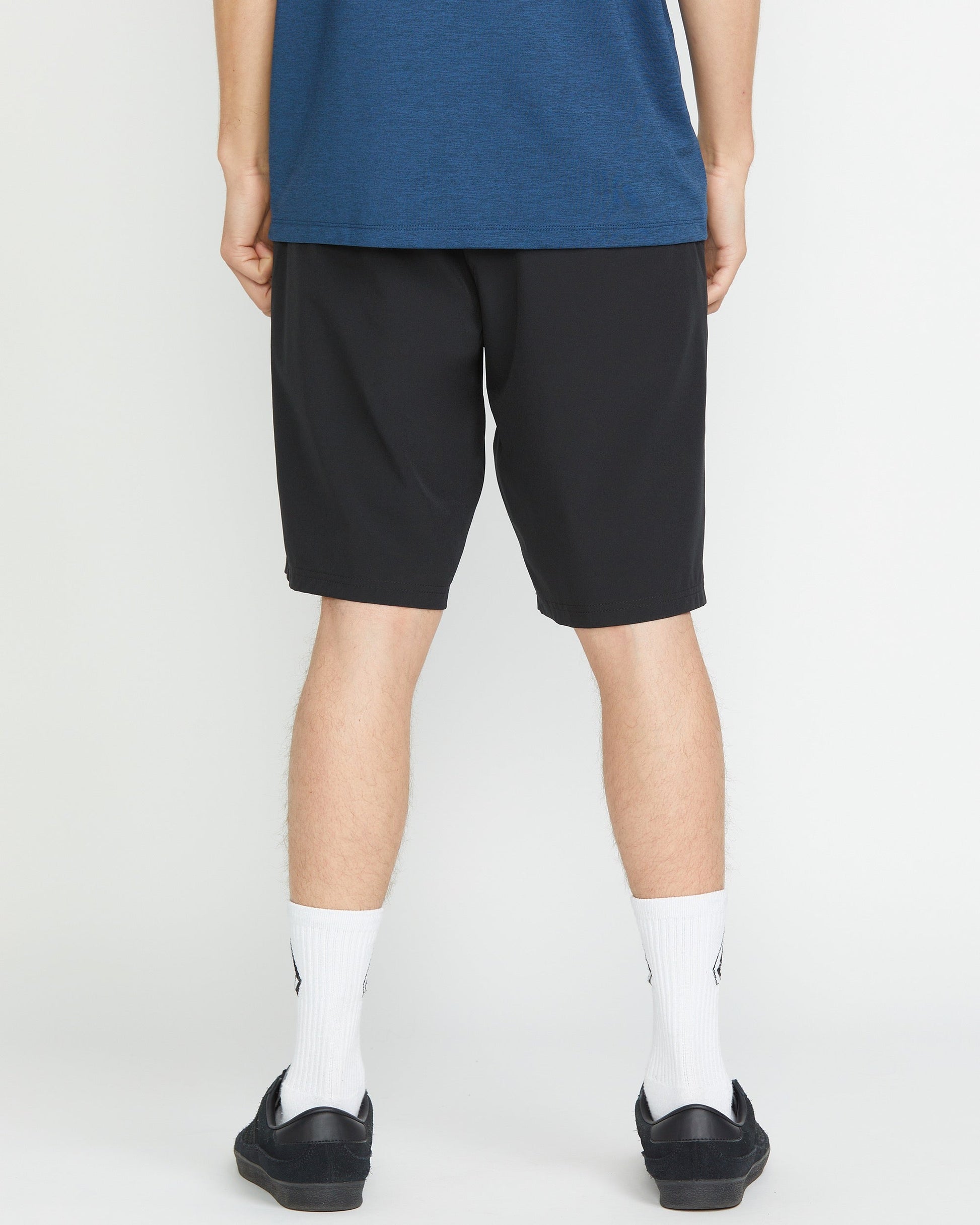 A person faces away from the camera, wearing a blue shirt and Volcom Men's Frickin Cross Shred 20" Shorts with white socks and black shoes, set against a plain white background.