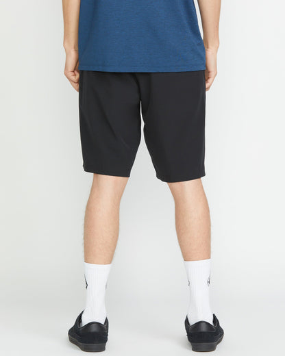 A person faces away from the camera, wearing a blue shirt and Volcom Men's Frickin Cross Shred 20" Shorts with white socks and black shoes, set against a plain white background.