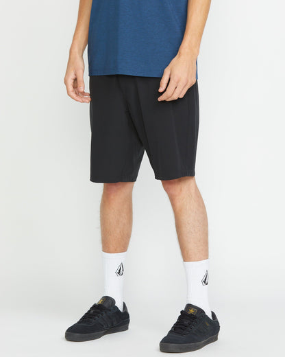 A person is seen from the shoulders down, wearing Volcom Men's Frickin Cross Shred 20" Shorts in blue, white crew socks with a black logo, and black sneakers, standing against a white background.