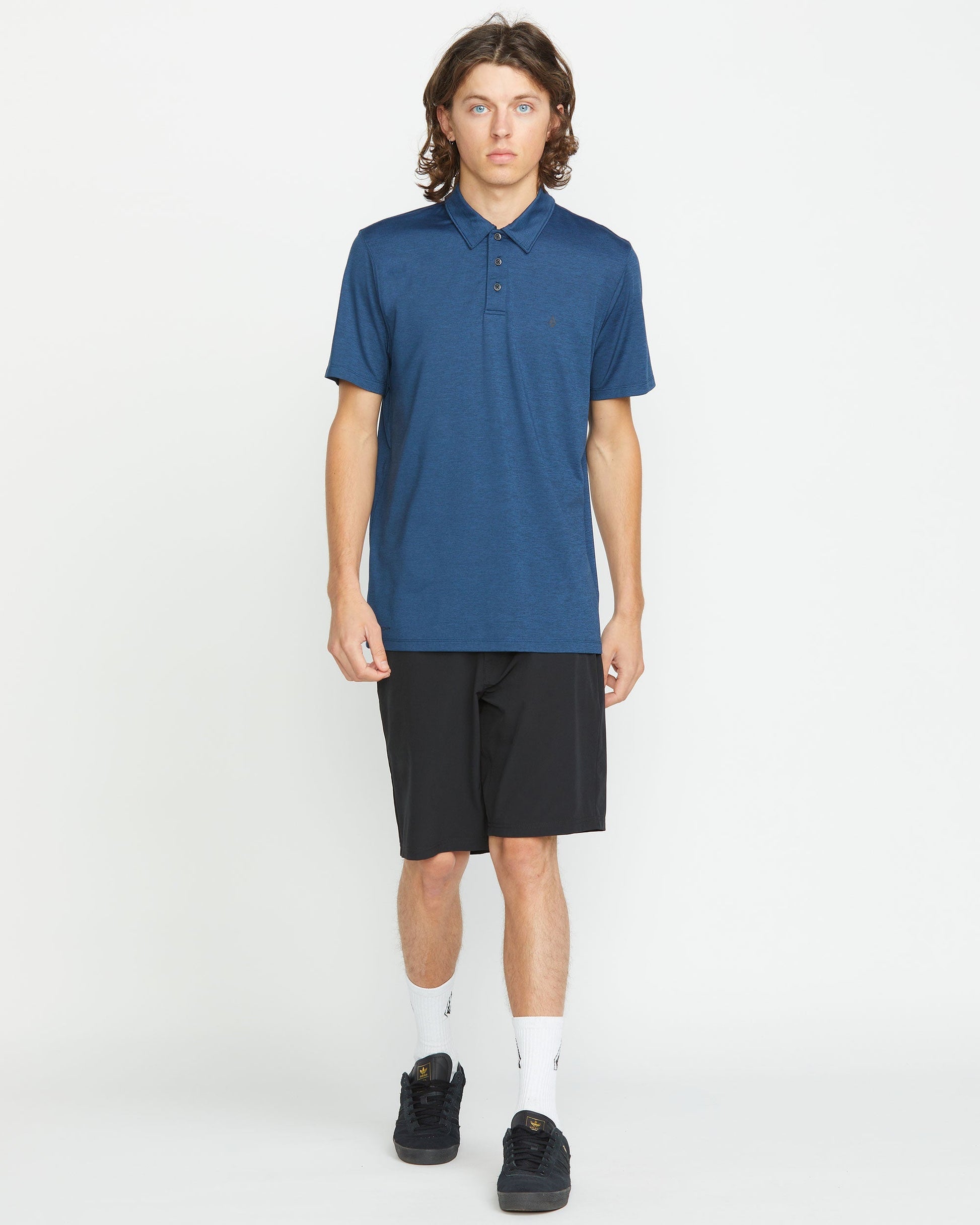 A person with long brown hair wears a dark blue polo and Volcom Men's Frickin Cross Shred 20" Shorts, paired with white socks and black shoes, standing against a plain white background.