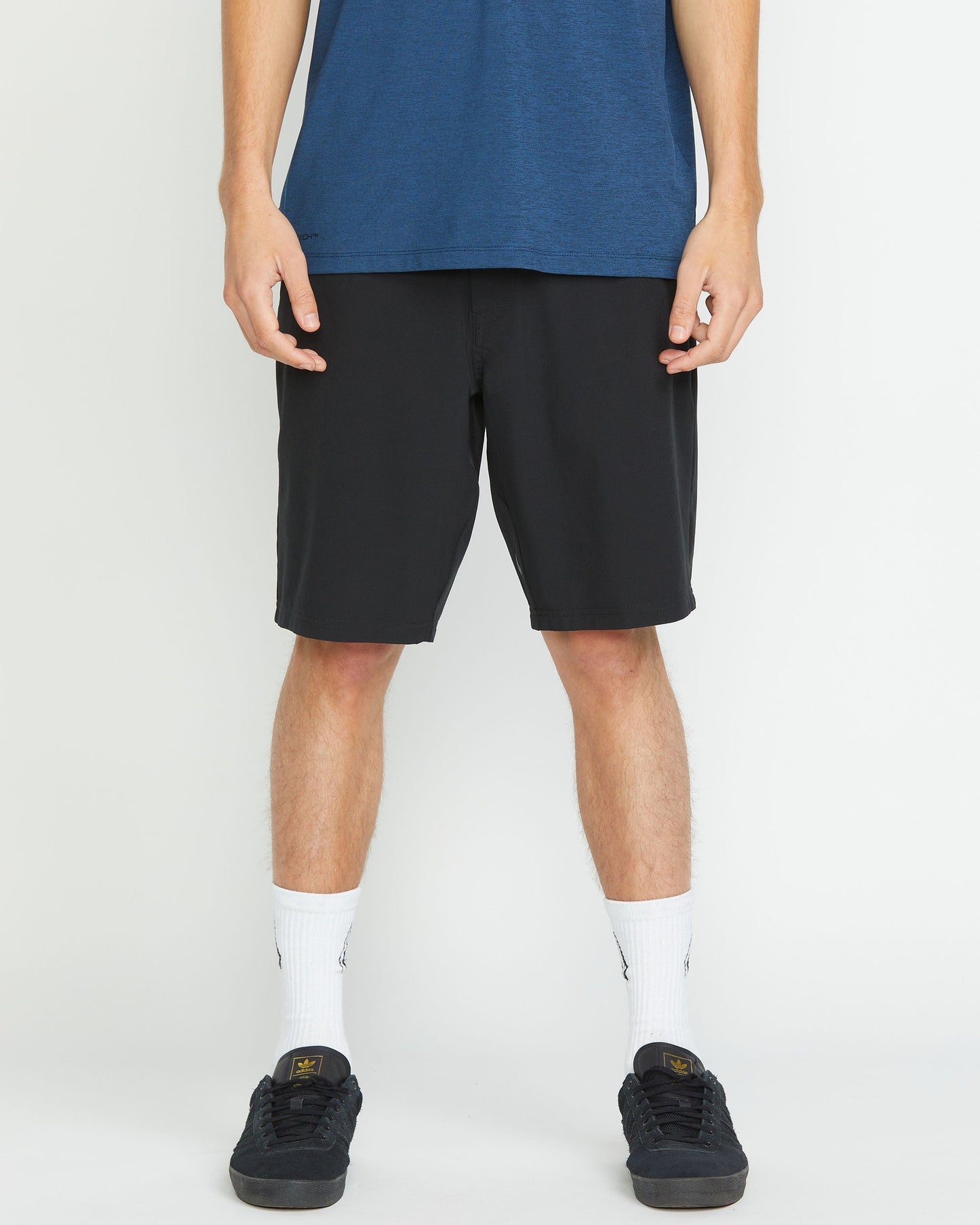 A person stands against a white background wearing a blue shirt, white crew socks, black sneakers, and Volcom Men's Frickin Cross Shred 20" Shorts in black. The image shows the person from shoulders to feet.