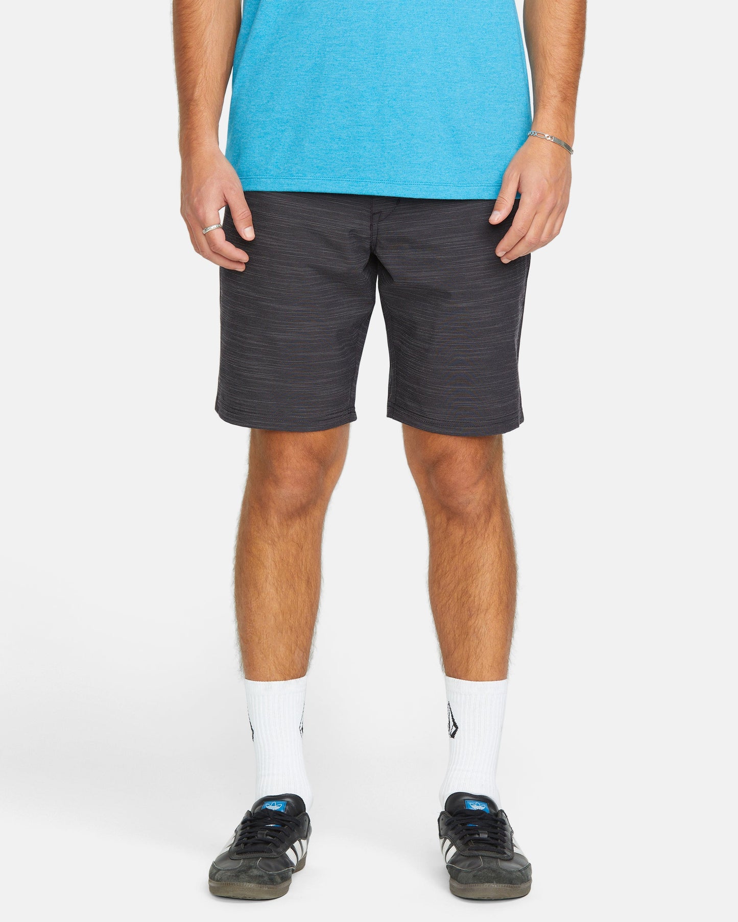 Only the lower half of a person wearing Volcom Men's Slub Frickin Cross Shred 20" Hybrid Shorts is visible as they stand facing forward against a plain white background, paired with white crew socks and black sneakers.