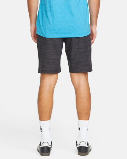 A person faces away, wearing a blue shirt, Volcom Men's Slub Frickin Cross Shred 20" Hybrid Shorts, white socks with a black logo, and black slide sandals against a plain white background.