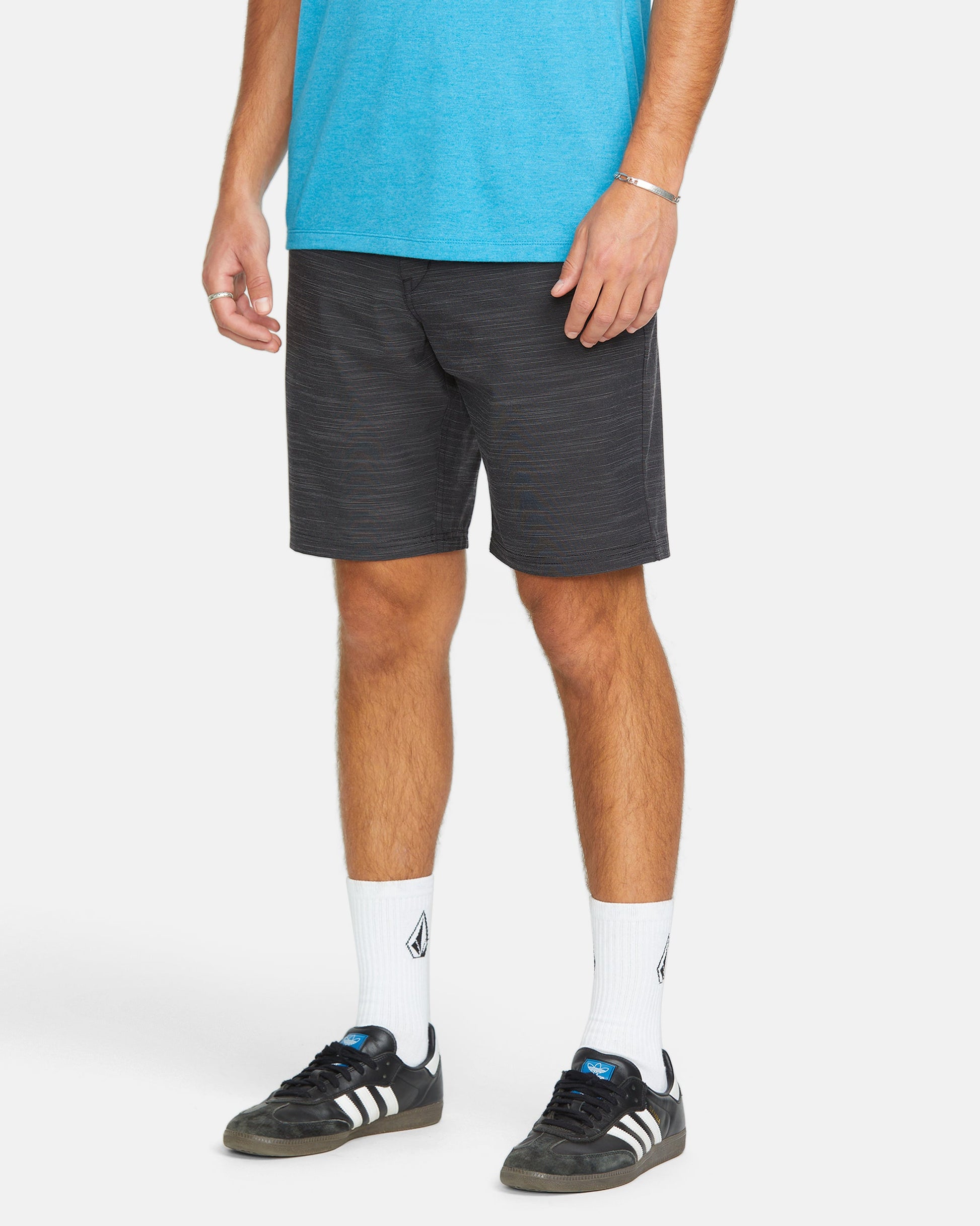 A person wears a blue T-shirt, Volcom Men's Slub Frickin Cross Shred 20" Hybrid Shorts, white socks with a black logo, and black Adidas sneakers with white stripes, standing against a white background.