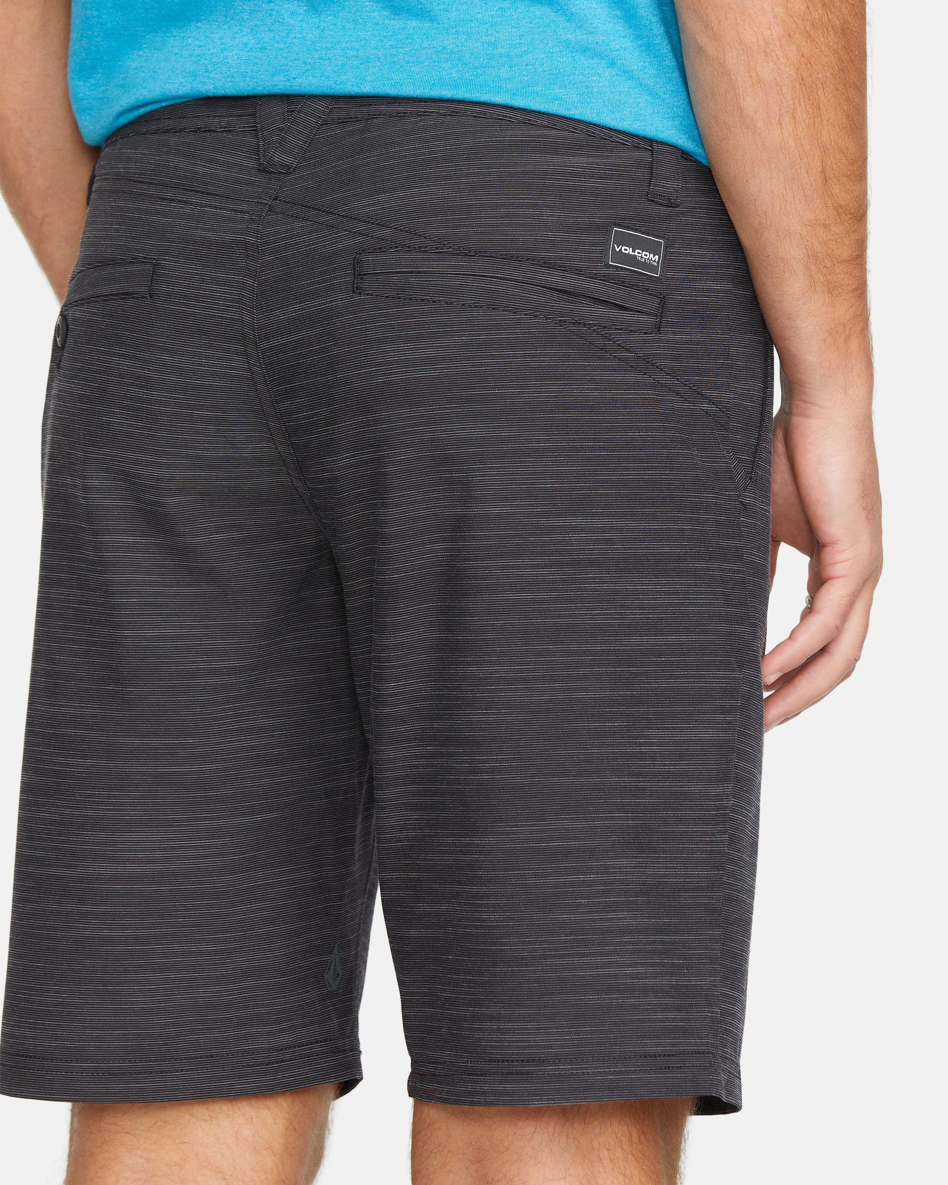 A person wears dark gray Volcom Men's Slub Frickin Cross Shred 20" Hybrid Shorts with 4-way stretch and a blue shirt, standing with their left hand down. A small rectangular Volcom logo patch is on the back right pocket.