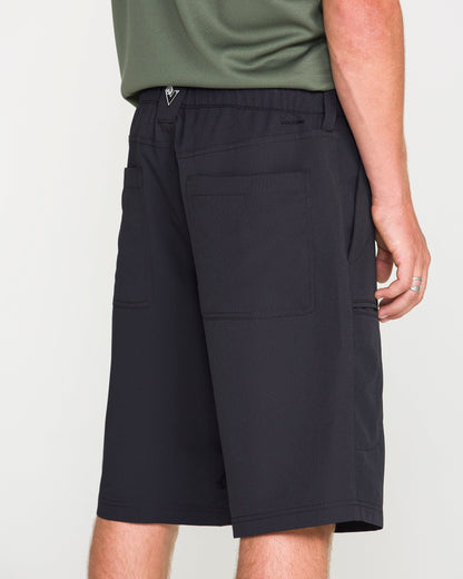 A person wearing Volcom Men's Venture Stone Hybrid Shorts in Asphalt Black, featuring knee-length recycled-poly fabric and large back pockets, along with a green shirt and a silver ring, standing against a plain white background.
