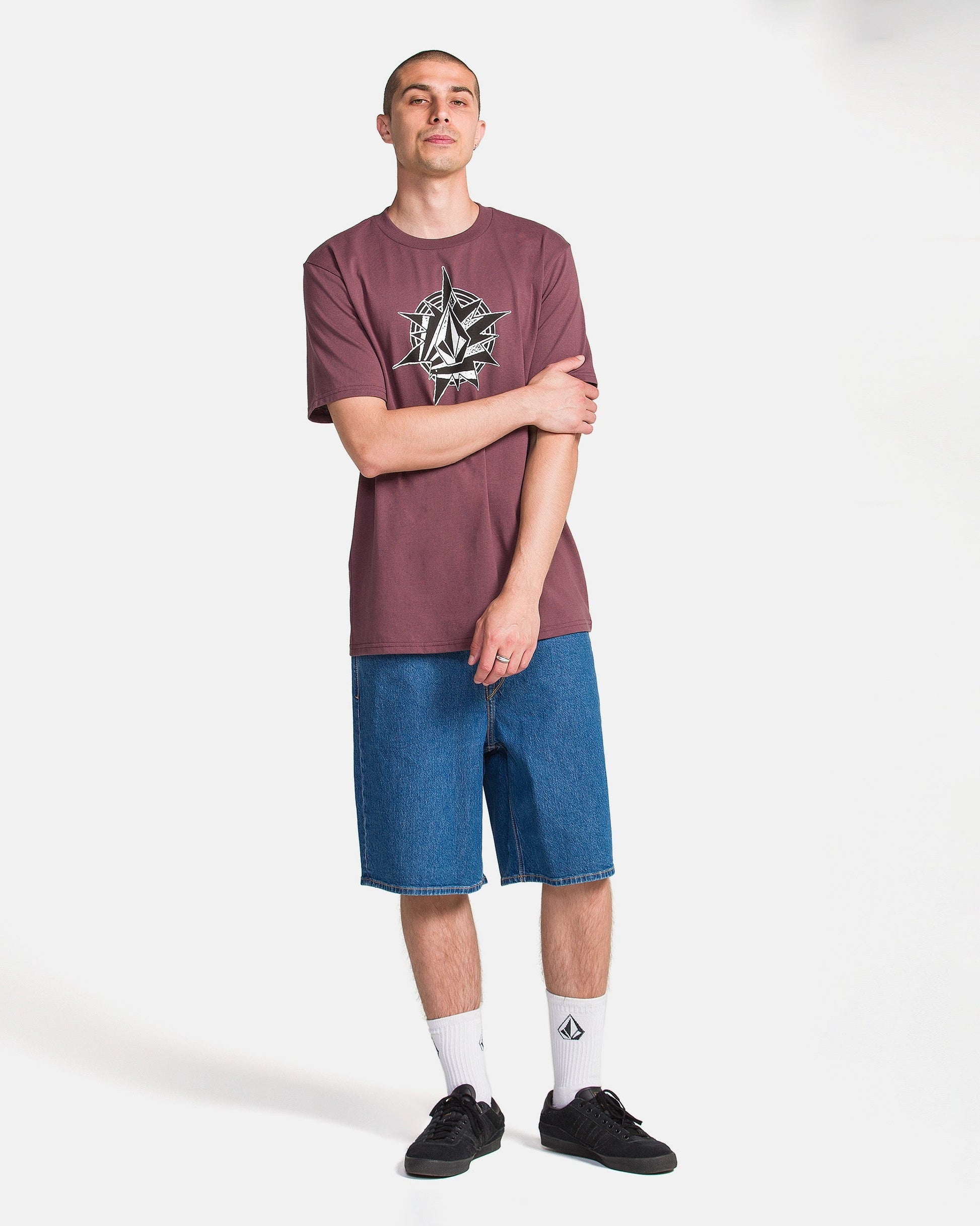 Against a white background, a person with short hair wears the Volcom Men's Yoink Short Sleeve Tee in maroon (100% cotton, classic fit, crew neck), blue denim shorts, white logo socks, and black sneakers. One hand touches their opposite arm.