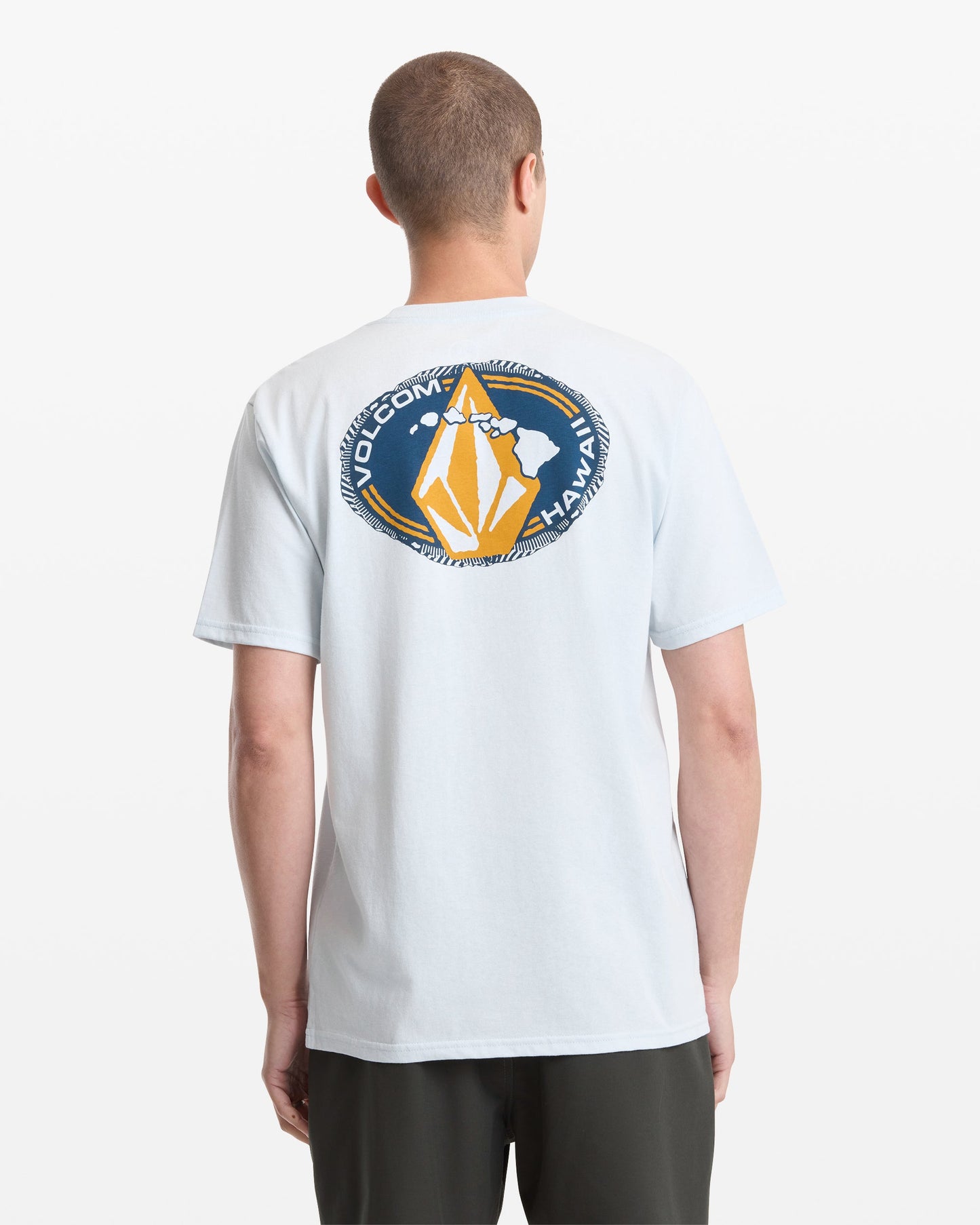 A person viewed from behind wears the Volcom Men's Loopy Short Sleeve Tee in light blue, featuring a circular graphic with "VOLCOM HAWAII" and a stylized yellow and white design on the back.