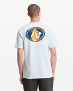 A person viewed from behind wears the Volcom Men's Loopy Short Sleeve Tee in light blue, featuring a circular graphic with "VOLCOM HAWAII" and a stylized yellow and white design on the back.