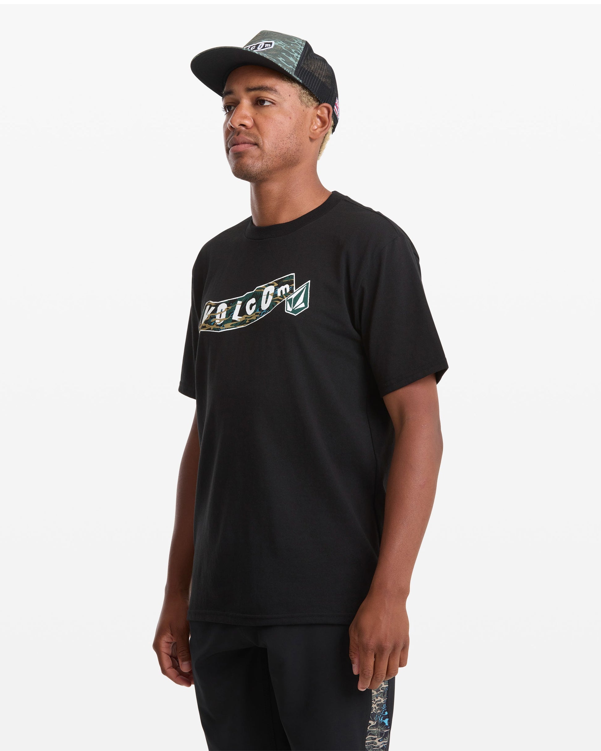 A man wearing a Volcom Men's Featured Artist Parillo Short Sleeve Tee, a black mesh cap, and black shorts stands against a plain white background, looking slightly to his left.