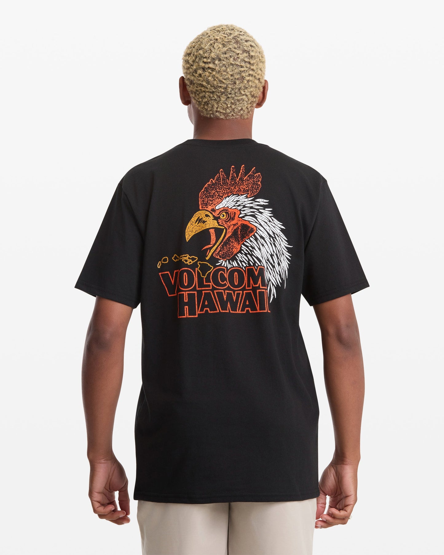 A person with short blond hair is seen from behind wearing the Volcom Men's Yewster Rooster Short Sleeve Tee, a black T-shirt with a colorful rooster graphic and "VOLCOM HAWAII" text on the back.