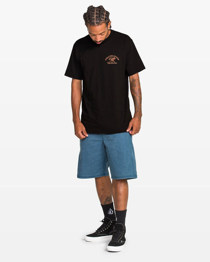 A man with braided hair looks down, wearing the Volcom Men's Crosscut Workwear Short Sleeve Tee for all-cotton comfort, paired with blue knee-length shorts, black socks with a white logo, and black sneakers against a plain white background.