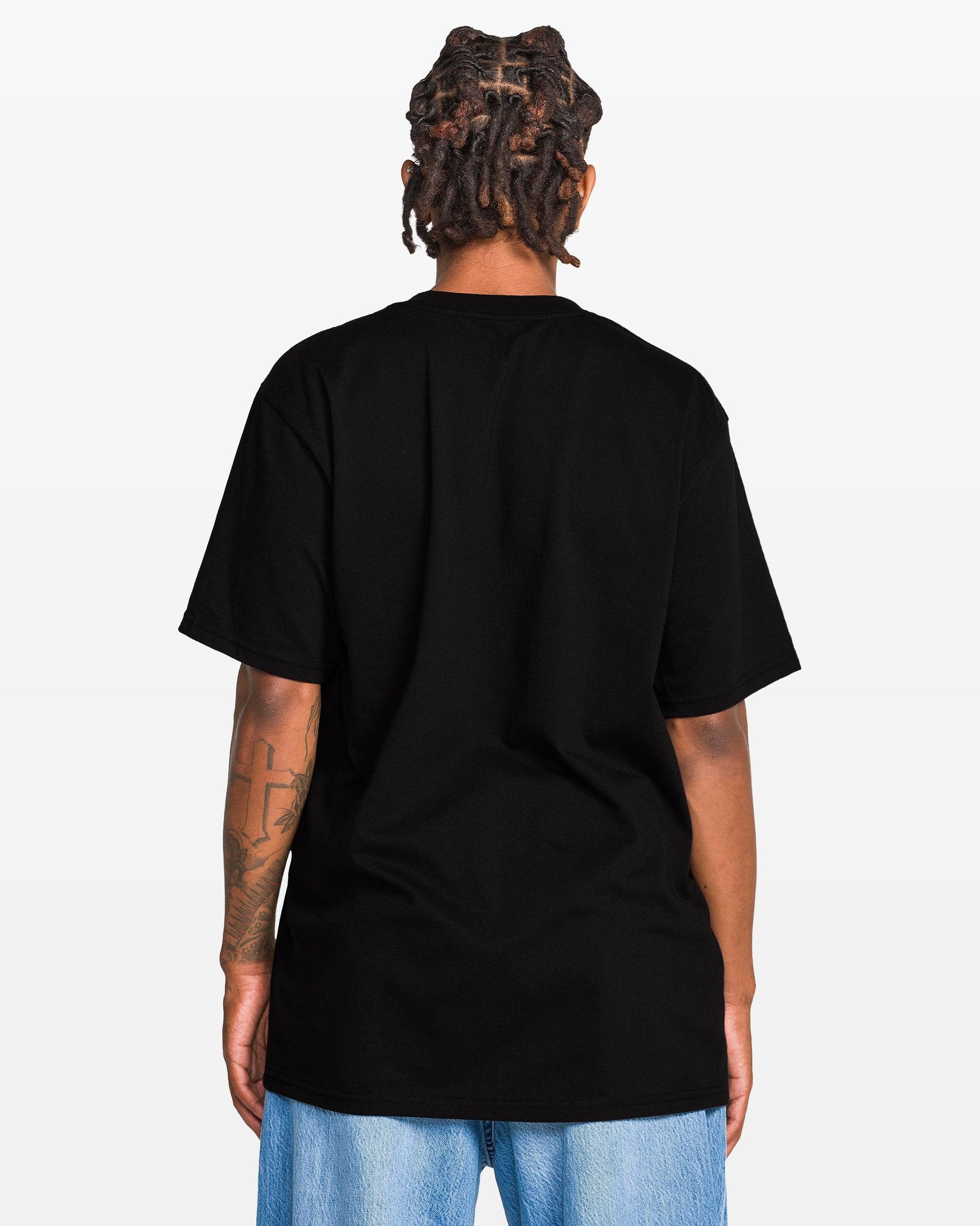 Person with short dreadlocks, seen from the back in a plain black Volcom Men's Eurobox Short Sleeve Tee (100% cotton) and light blue jeans, against a white background. Tattoo is visible on left arm.