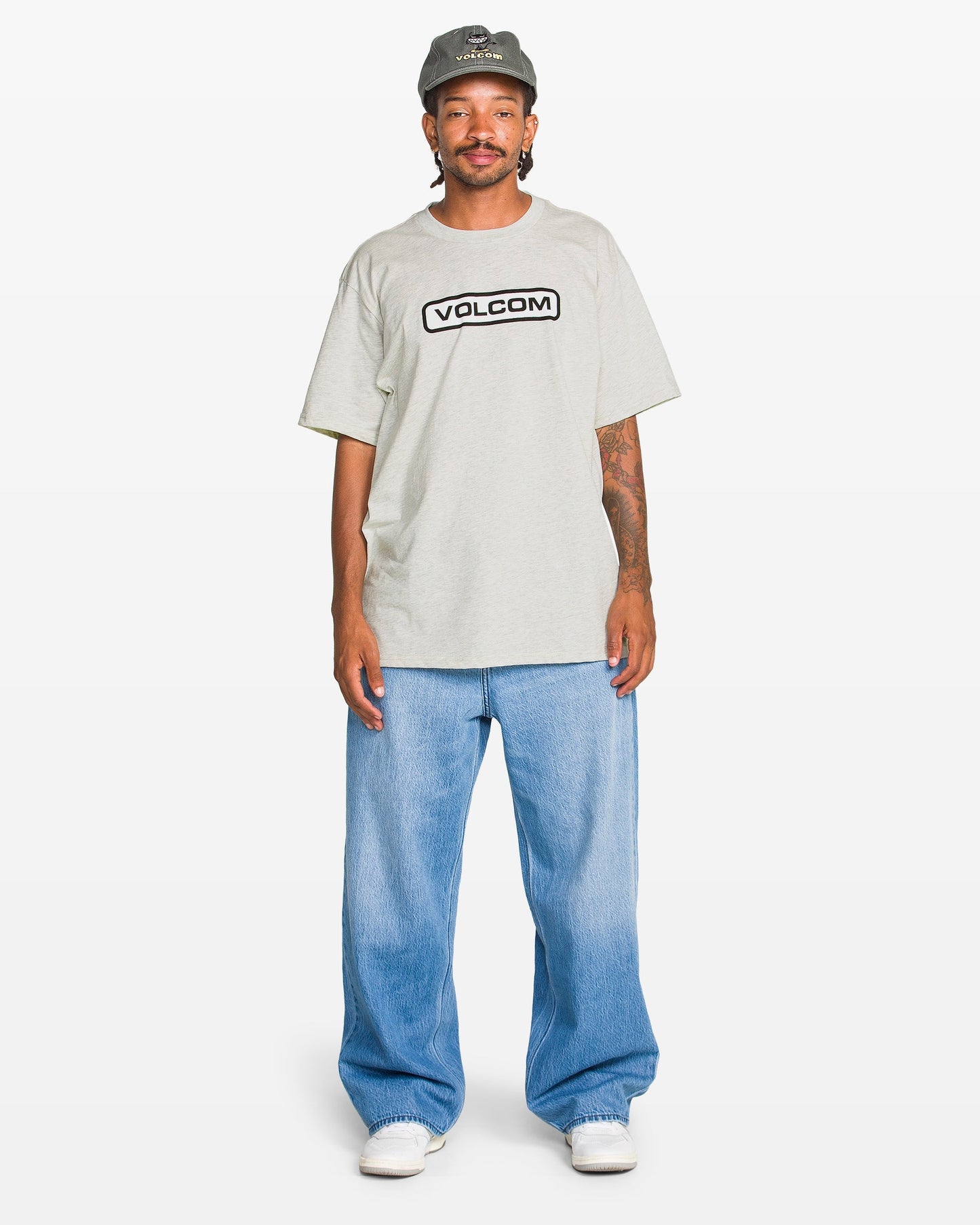 A person wears a gray Volcom Men's Eurobox Short Sleeve Tee with a soft-hand print, light blue baggy jeans, white sneakers, and a gray cap while standing against a plain white background.