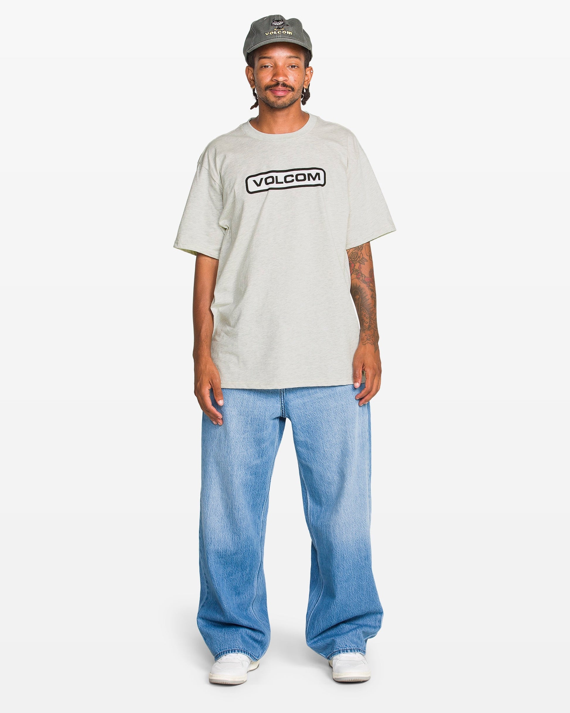 A person wears a gray Volcom Men's Eurobox Short Sleeve Tee with a soft-hand print, light blue baggy jeans, white sneakers, and a gray cap while standing against a plain white background.