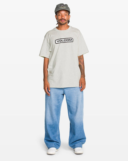 A person wears a gray Volcom Men's Eurobox Short Sleeve Tee with a soft-hand print, light blue baggy jeans, white sneakers, and a gray cap while standing against a plain white background.