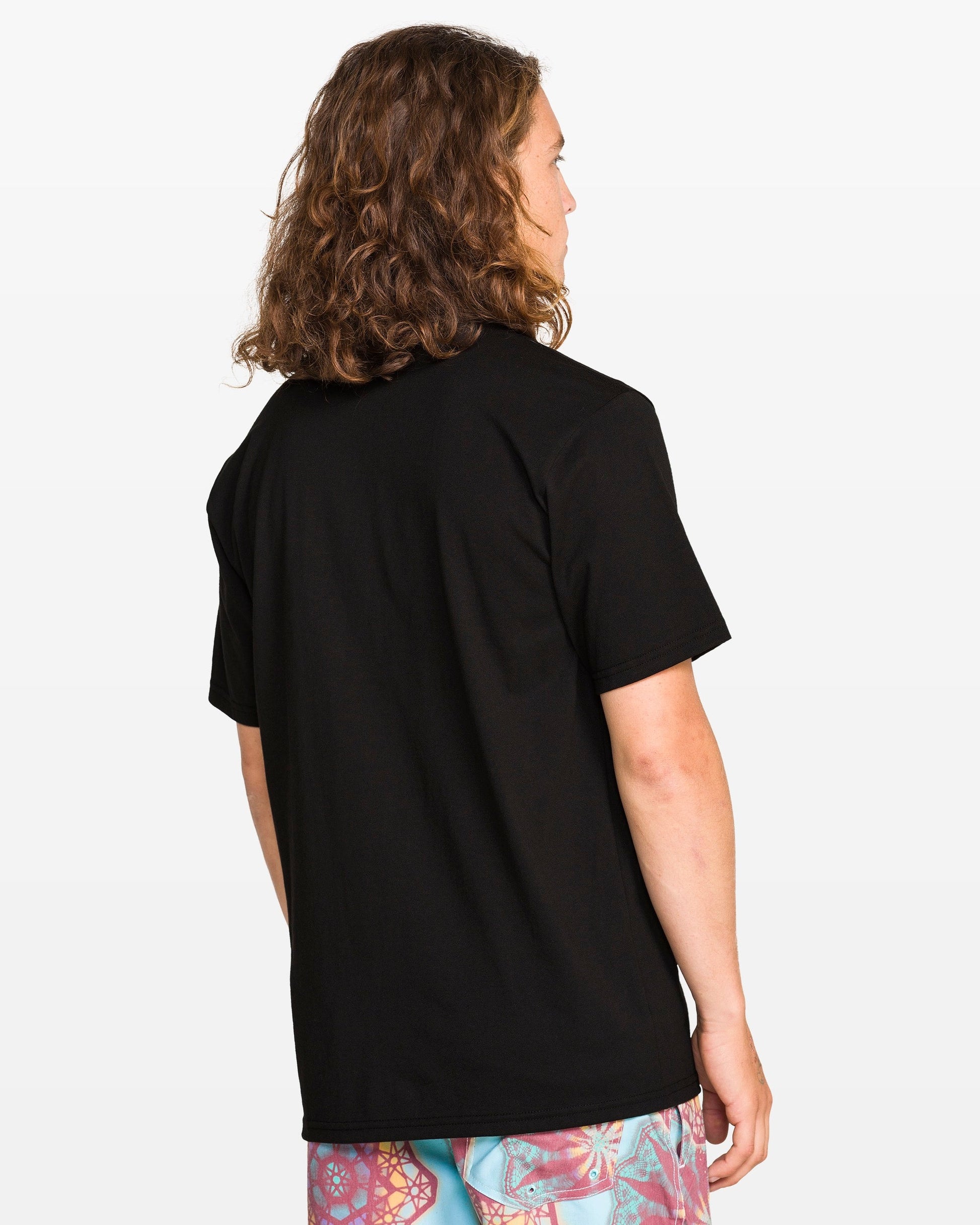 A person with long curly hair stands with their back to the camera, wearing the Volcom Men's Star Shields Featured Artist Exp Short Sleeve Tee and colorful patterned shorts against a white background.