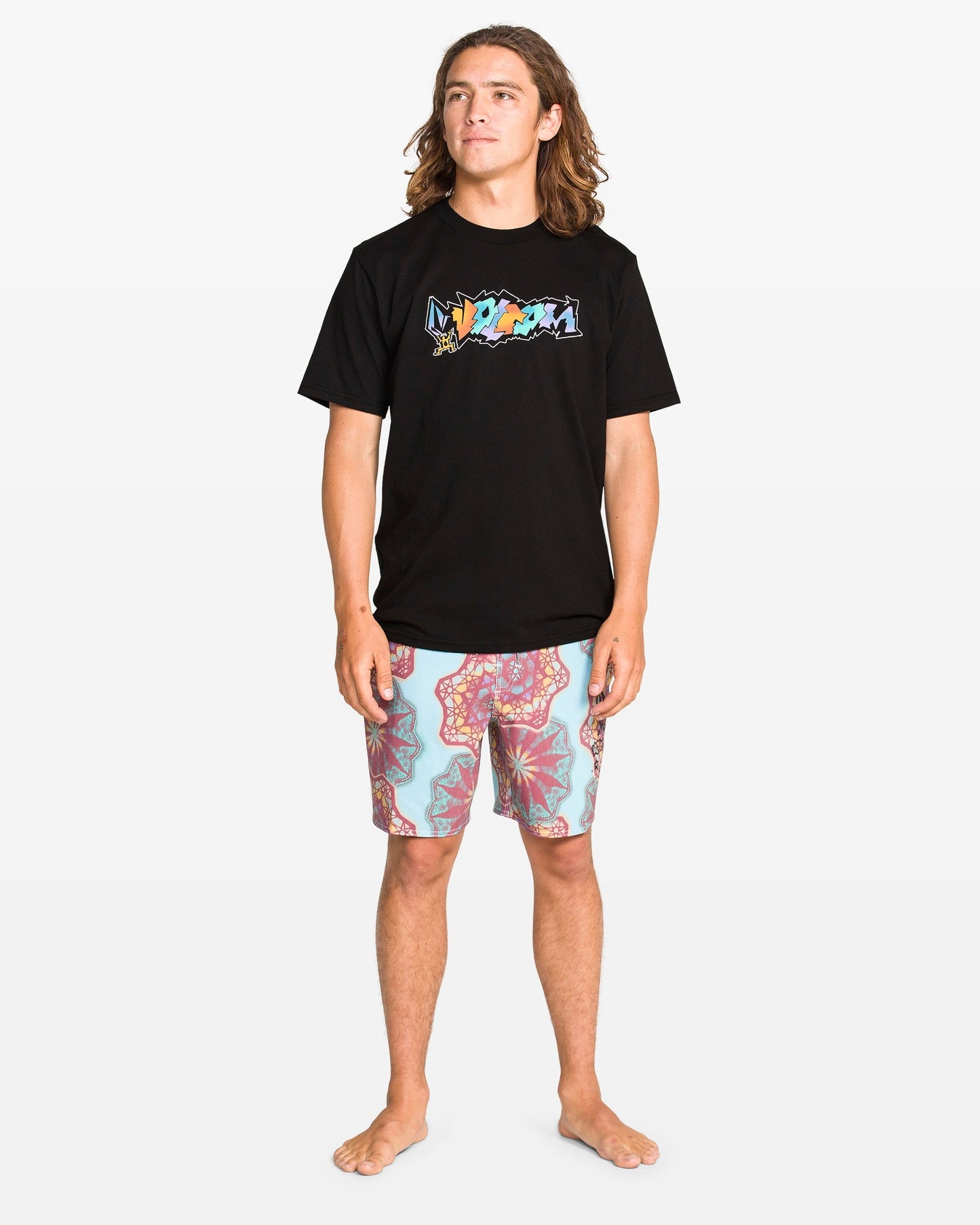 A young man with long hair stands barefoot, wearing a Volcom Men's Star Shields Featured Artist Exp Short Sleeve Tee and patterned pink and blue shorts, set against a plain white background.