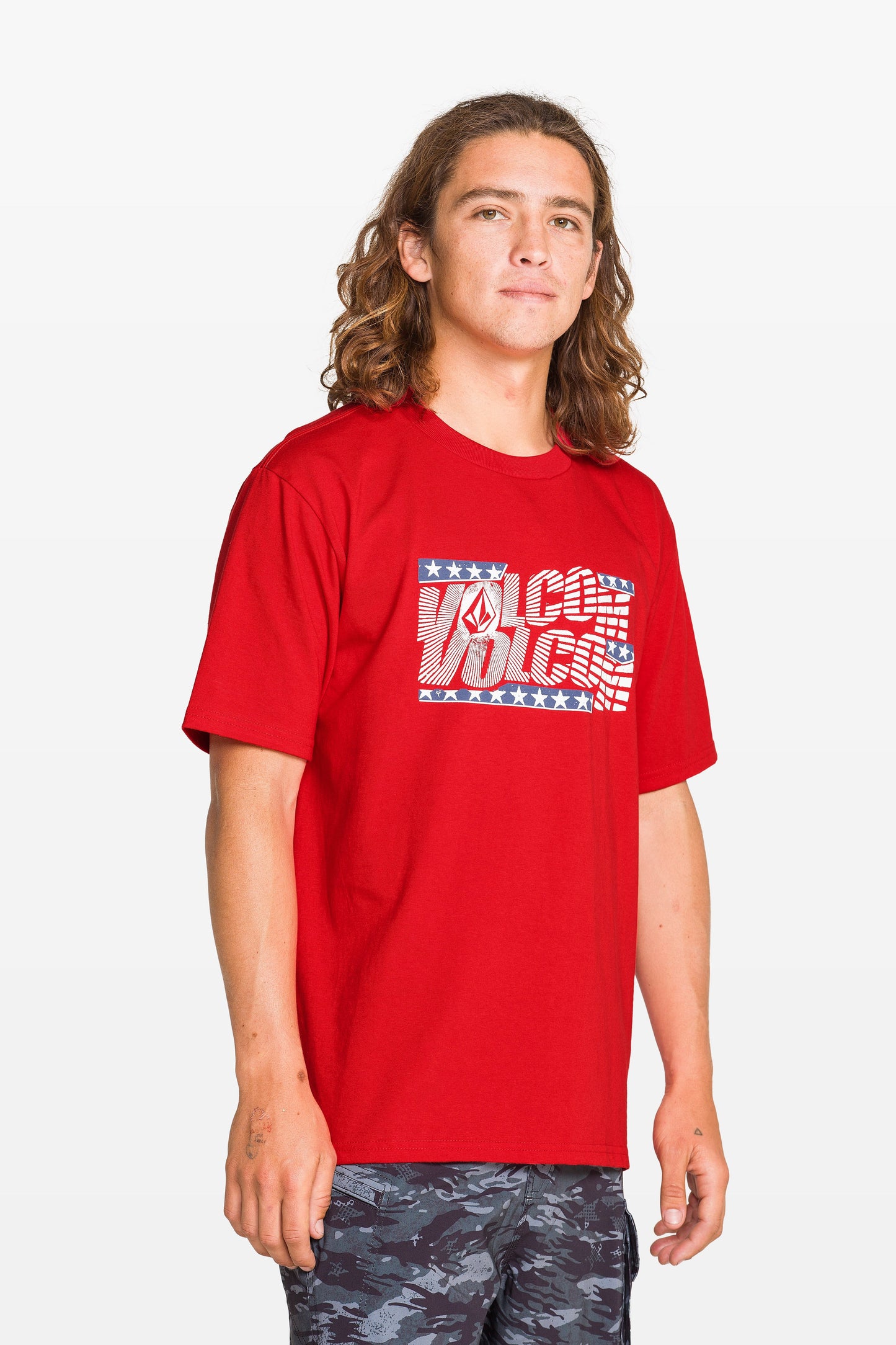 A child with long brown hair wears a Kids' Glory Daze Short Sleeve Tee by Volcom, made from 100% cotton with a patterned logo. They stand against a light background, looking relaxed and wearing dark patterned shorts.