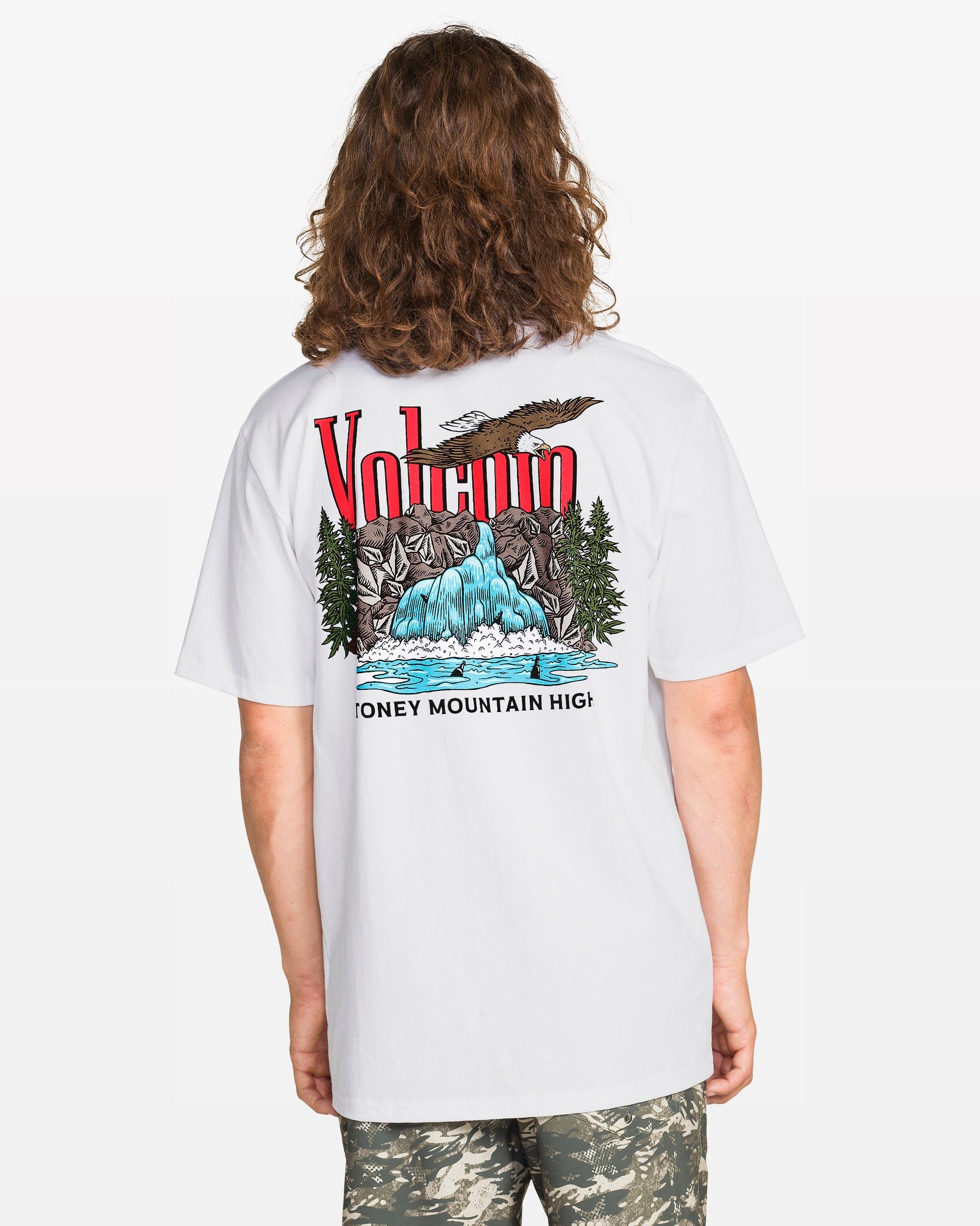 A person with long hair wears the Volcom Men's Stoney Mountain Short Sleeve Tee, a classic fit 100% cotton shirt with a colorful mountain and waterfall graphic on the back, paired with camouflage shorts.