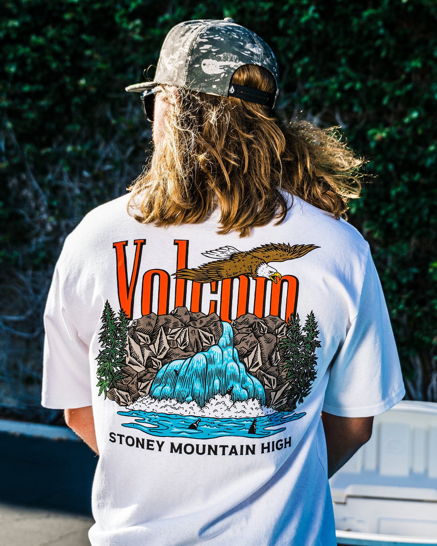 A person with long hair wears a camouflage cap and the Volcom Men's Stoney Mountain Short Sleeve Tee, made from 100% cotton with a vibrant waterfall, mountains, eagle, and trees graphic, plus VOLCANO and STONEY MOUNTAIN HIGH on the back.