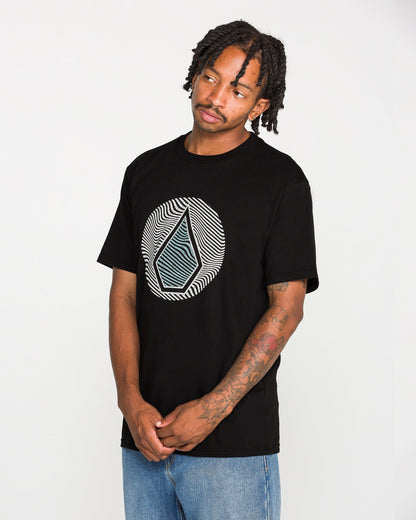 A man with braided hair, a mustache, and tattoos on his forearm wears the Volcom Men's Wave Stone Short Sleeve Tee—a classic fit t-shirt made from premium ring spun cotton and featuring a geometric design. He pairs it with blue jeans and stands against a plain white background.