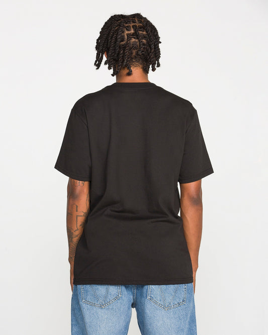 A person with short, twisted hair stands facing away, wearing the Volcom Men's Wave Stone Short Sleeve Tee in plain black, crafted from premium ring-spun cotton in a classic fit, paired with blue jeans. Their arms are visible, showcasing tattoos on the left arm. The background is plain white.