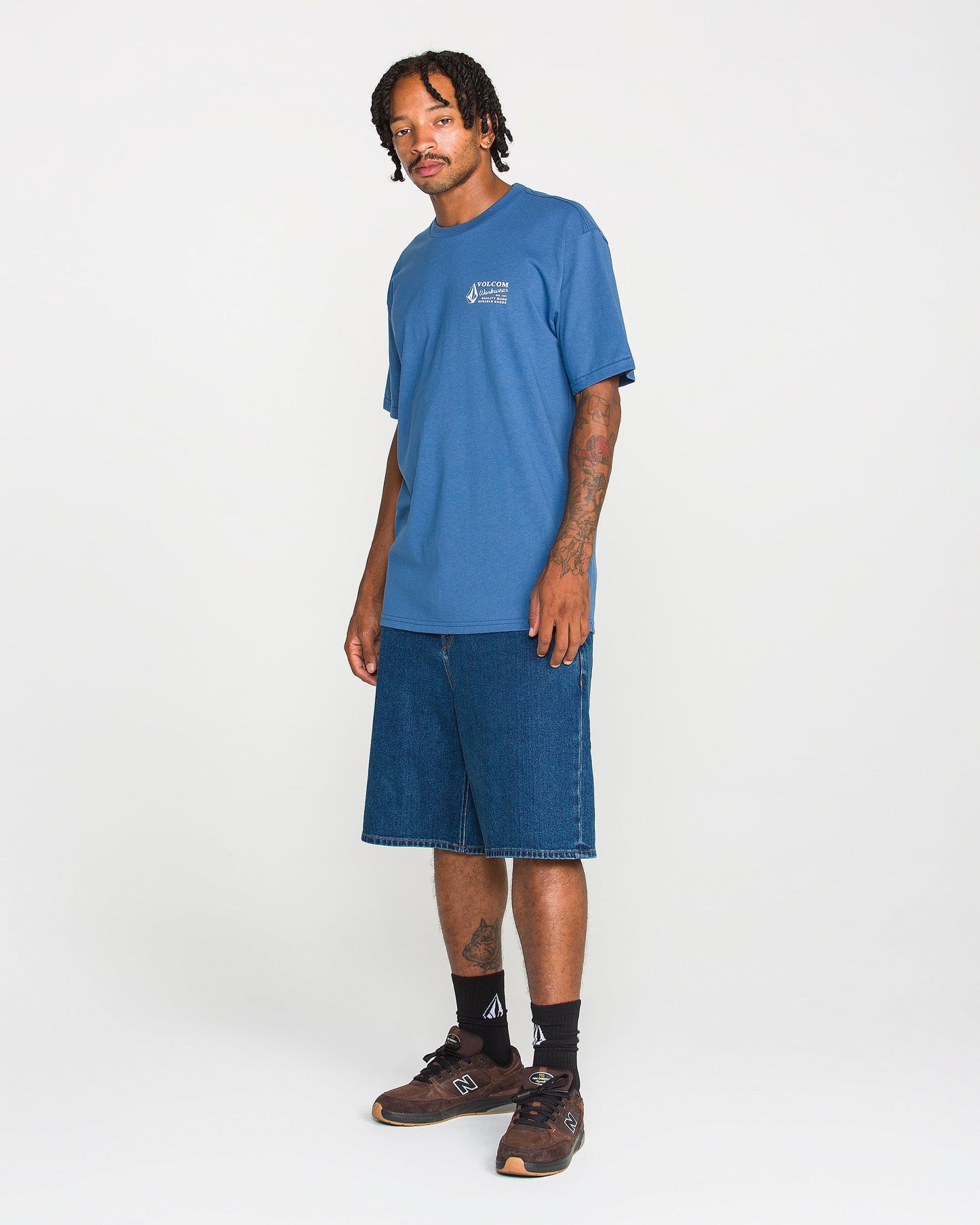 A man with braided hair wears the Men's Volcom Workwear Short Sleeve Tee in blue, styled with long denim shorts, black socks, and brown sneakers as he stands against a plain white background—a classic Volcom look.