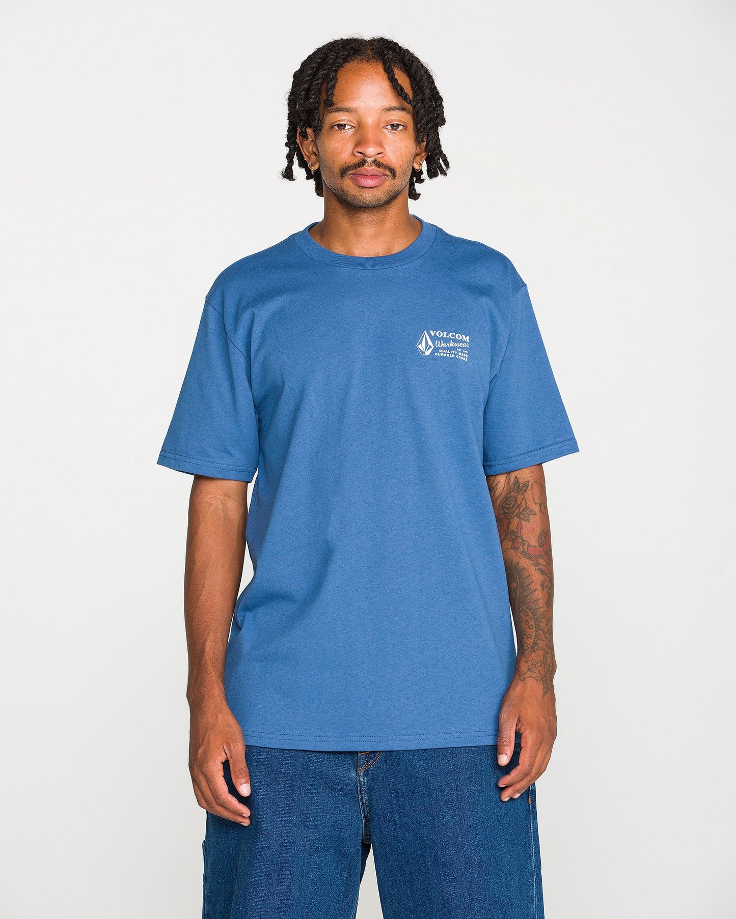 A young Black man with braided hair and a mustache stands against a white background, wearing the Volcom Men's Volcom Workwear Short Sleeve Tee in blue paired with blue jeans. He has visible tattoos on his left arm and is looking directly at the camera.