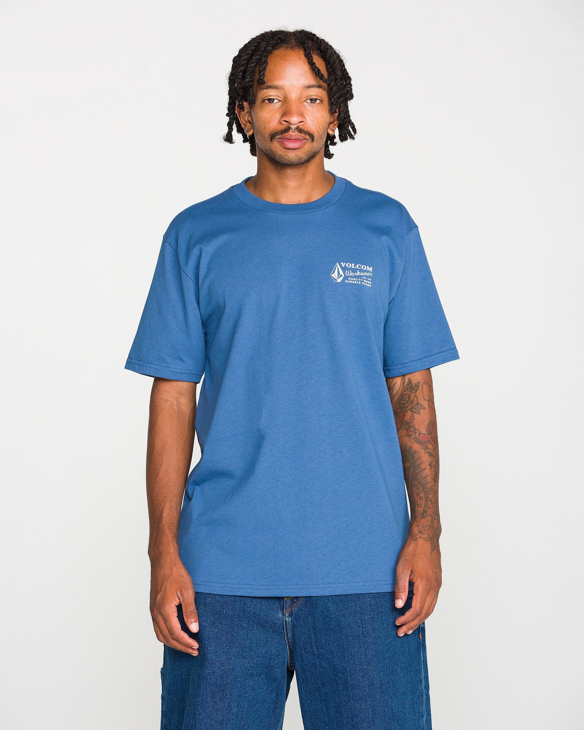 A young Black man with braided hair and a mustache stands against a white background, wearing the Volcom Men's Volcom Workwear Short Sleeve Tee in blue paired with blue jeans. He has visible tattoos on his left arm and is looking directly at the camera.