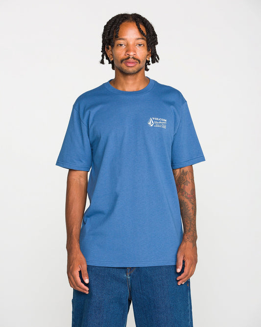 A young Black man with braided hair and a mustache stands against a white background, wearing the Volcom Men's Volcom Workwear Short Sleeve Tee in blue paired with blue jeans. He has visible tattoos on his left arm and is looking directly at the camera.