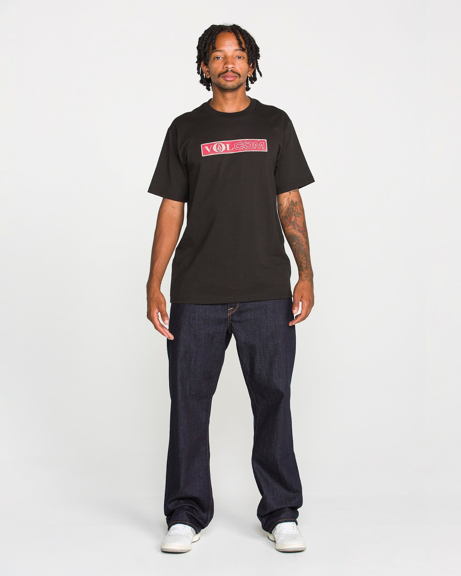 A man with short dreadlocks wears the Volcom Men's Dual Euro Short Sleeve Tee, crafted from premium ring-spun cotton and featuring a bold red and white vintage logo. He pairs the black T-shirt with dark jeans and white sneakers, standing facing forward against a plain white background.