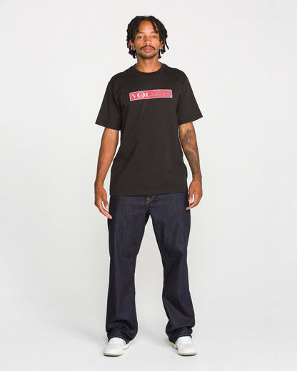A man with short dreadlocks wears the Volcom Men's Dual Euro Short Sleeve Tee, crafted from premium ring-spun cotton and featuring a bold red and white vintage logo. He pairs the black T-shirt with dark jeans and white sneakers, standing facing forward against a plain white background.