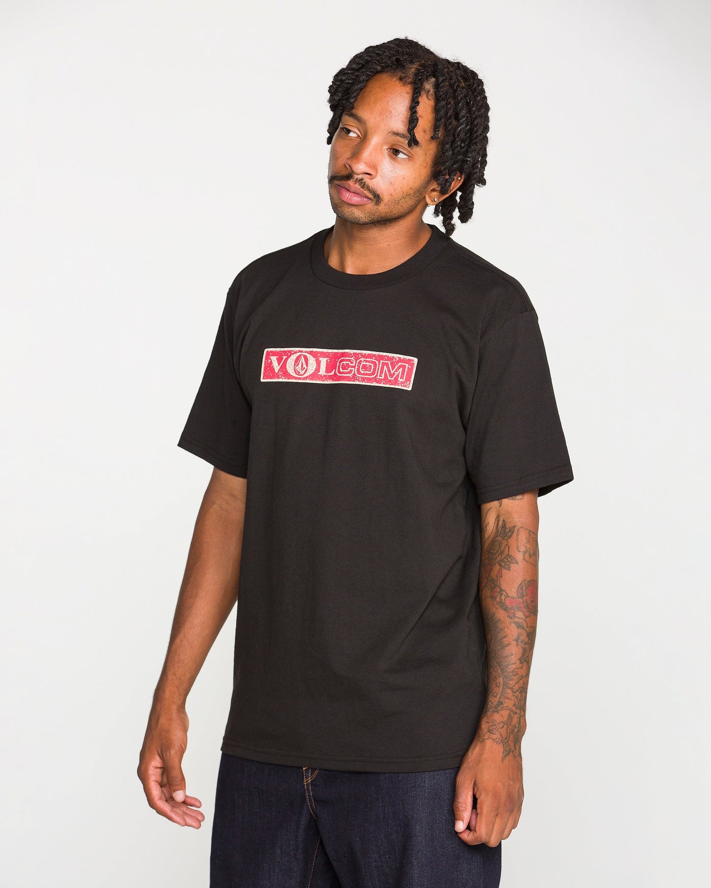 A man with short twists wears the Volcom Men's Dual Euro Short Sleeve Tee, a black t-shirt with a red and white vintage logo, made from premium ring-spun cotton, paired with dark jeans. He stands against a plain light background, looking slightly to his left.