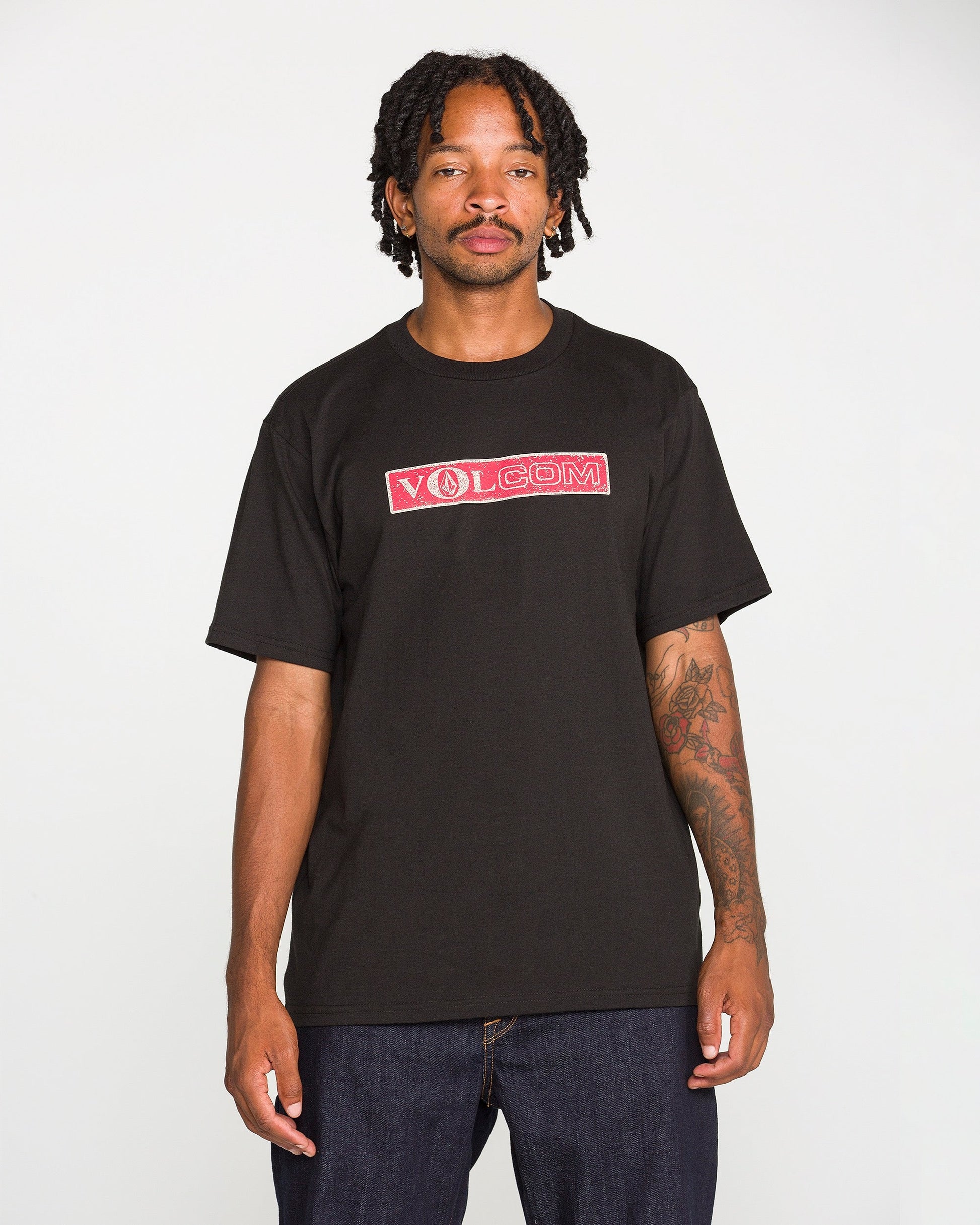 A man with tattoos and dreadlocks is wearing a Volcom Men's Dual Euro Short Sleeve Tee in black, made from premium ring-spun cotton, along with dark jeans. He stands against a plain white background, looking at the camera with a neutral expression.